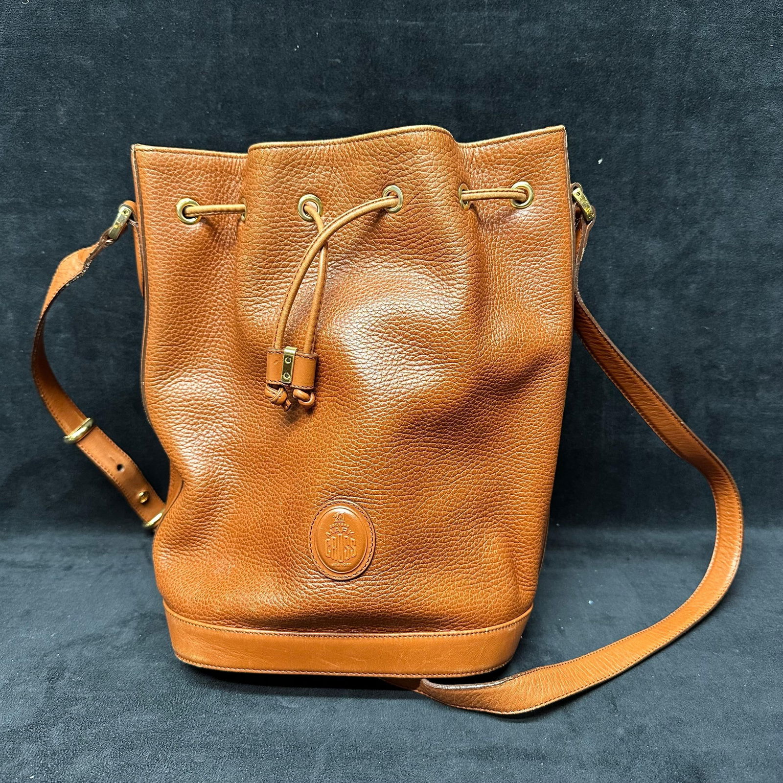 Mark Cross Brown Pebbled Leather Drawstring Bucket Bag: Mark Cross bucket-style handbag crafted in brown pebbled leather. Features a classic drawstring closure with brass-tone grommets and a leather tie, an adjustable shoulder strap, and a structured base.