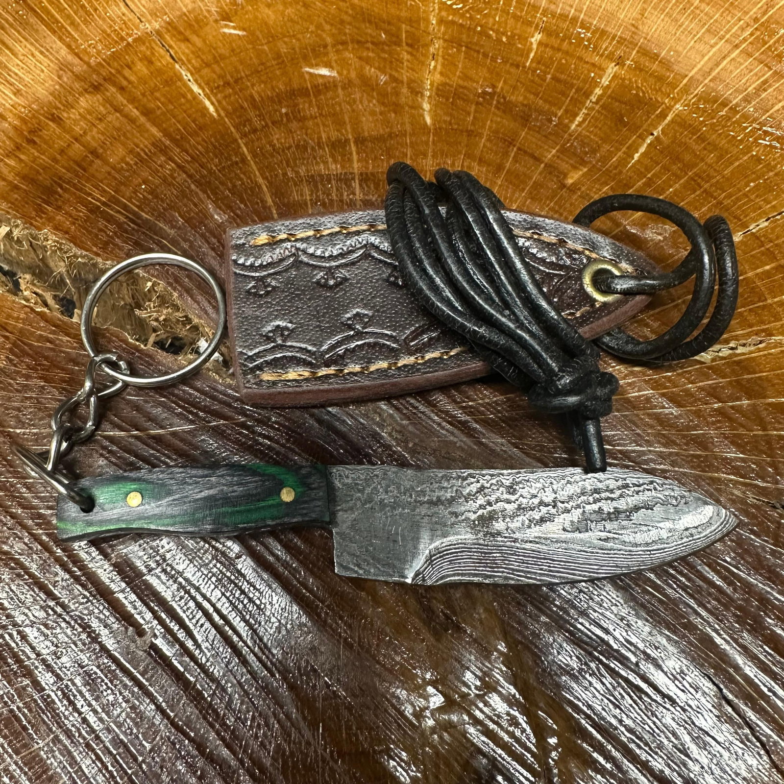 MHS Cutlery Keychain Butcher Style Blade Knife With Leather Case Necklace R44: Small key chain-sized fixed-blade knife featuring a patterned steel blade and a compact epoxy-resin/wood green/black-tone handle secured with brass pins. Includes a fitted brown leather sheath with st