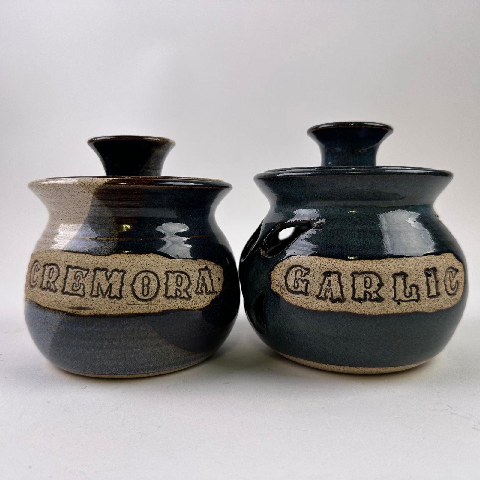 Handcrafted Pottery Crock Garlic and Cremora Jar Keeper Set Signed: Pair of handcrafted studio pottery jars labeled Garlic and Cremora. Blue and earth-tone glazed stoneware with matching lids. Both show signed maker's marks on the bottom base. No cracks observed.