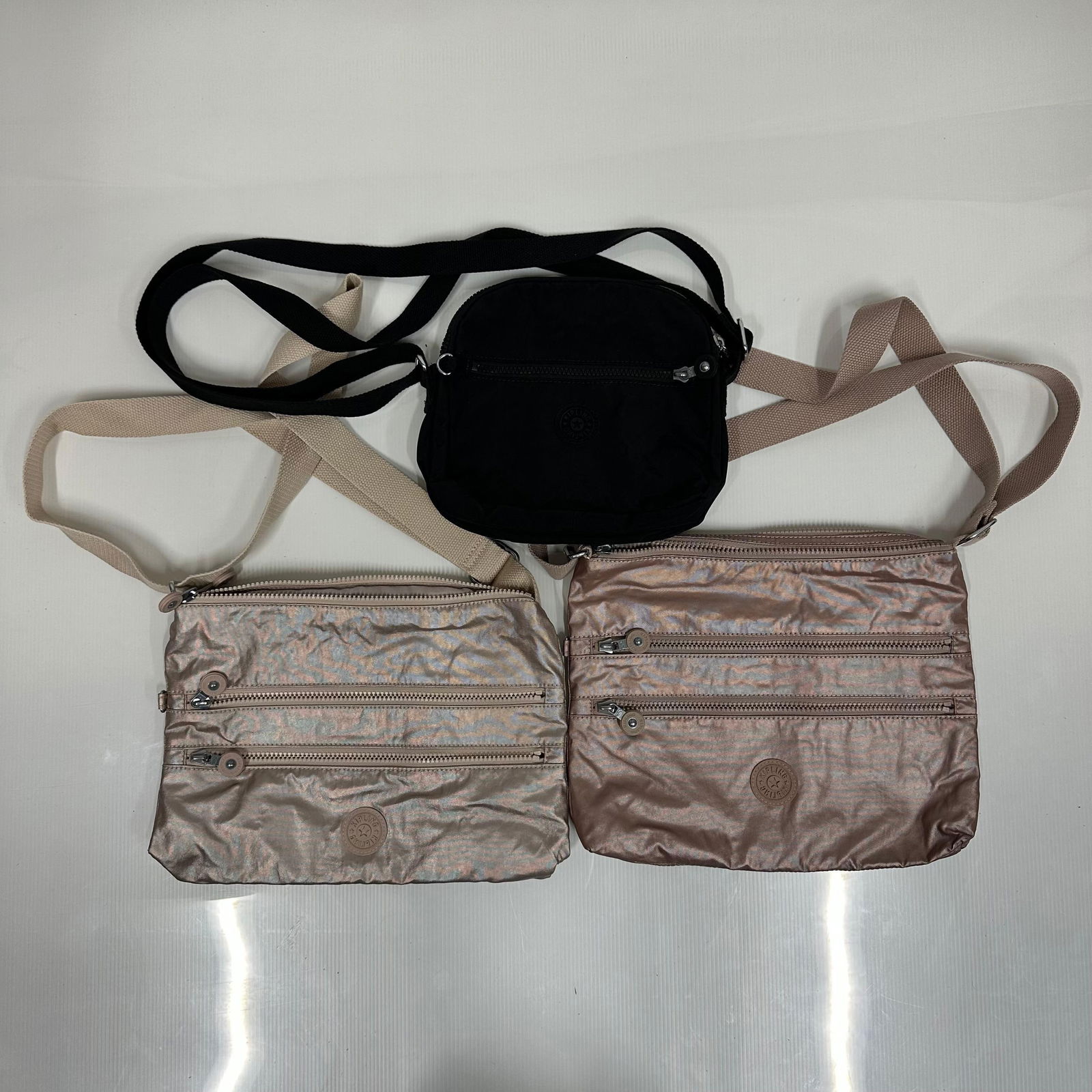 3 Kipling Crossbody Bags Two Metallic Rose Gold and One Black Nylon: Two Metallic rose gold finish crossbody bags with beige adjustable straps. Each features dual front zipper pockets, a top zipper closure, and a Kipling logo patch on the front. Interior includes a