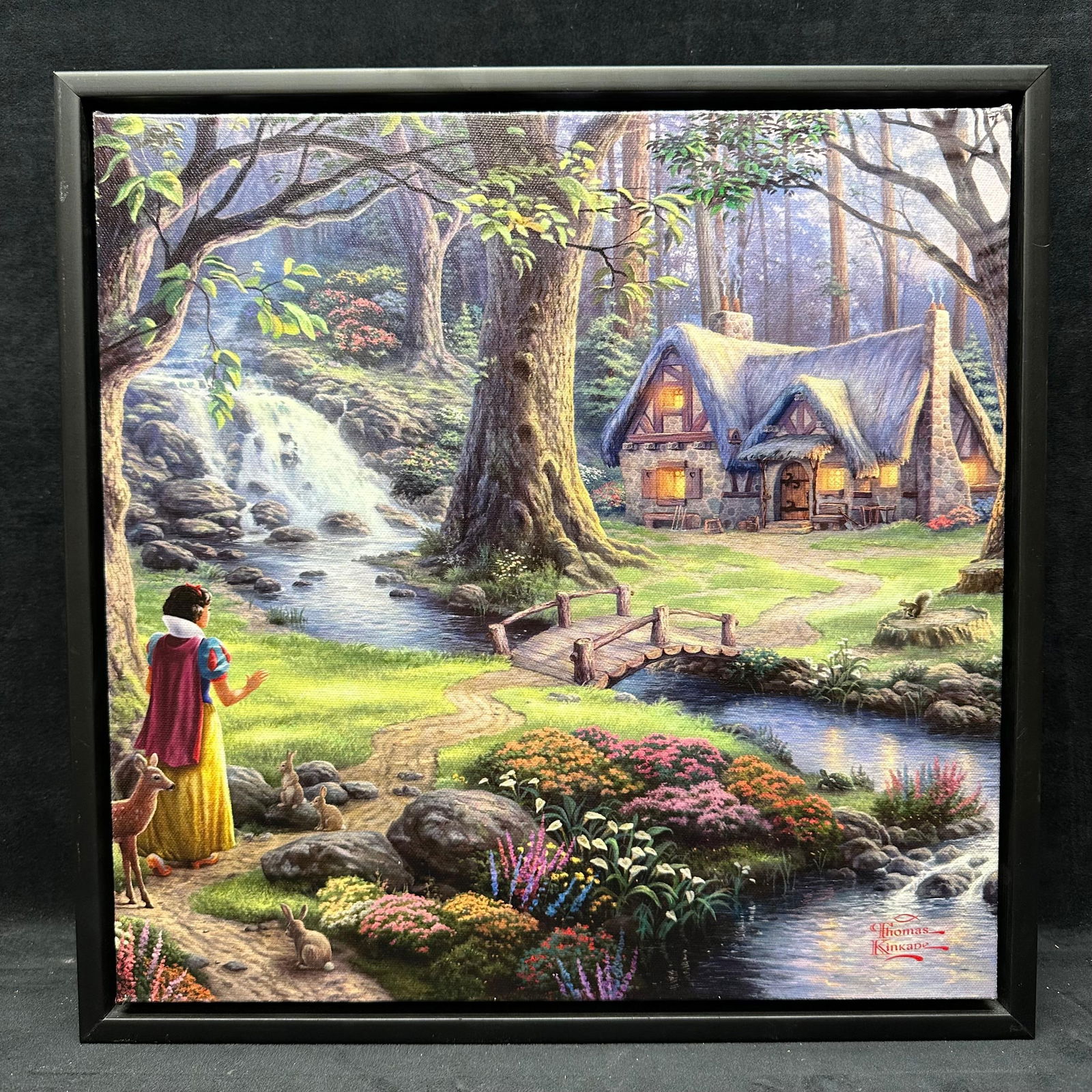 Thomas Kinkade Snow White Cottage Framed Accent Canvas Print: Framed accent canvas print after Thomas Kinkade, depicting a fairy tale woodland cottage scene inspired by Snow White. Printed canvas mounted on stretcher, presented in a black frame. Print signed in