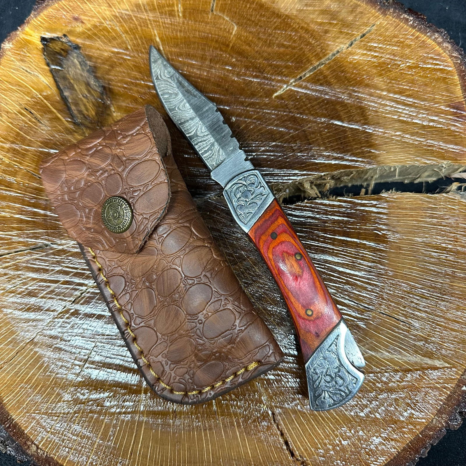 MHS Cutlery Steel Folding Knife with Wood Handle and Leather Sheath P49: MHS Cutlery folding knife featuring a patterned steel blade and a contoured wood handle with decorative metal bolsters. Includes a fitted brown leather sheath. Blade locks open; folding mechanism