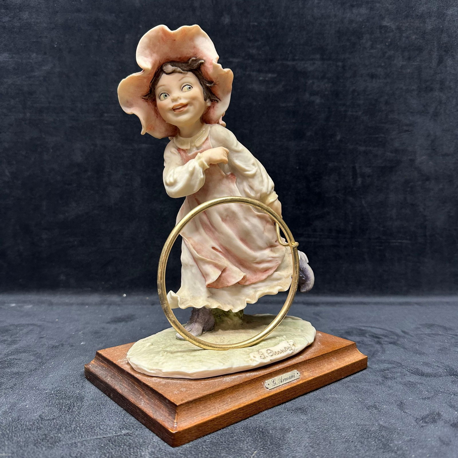 G. Armani Girl with Hoop Figural Sculpture Wooden Base: G. Armani sculpture depicting a young girl running with a brass hoop, wearing a flowing dress and a large bonnet. Hand-finished composition with soft pastel tones, mounted on a wooden display base. Si