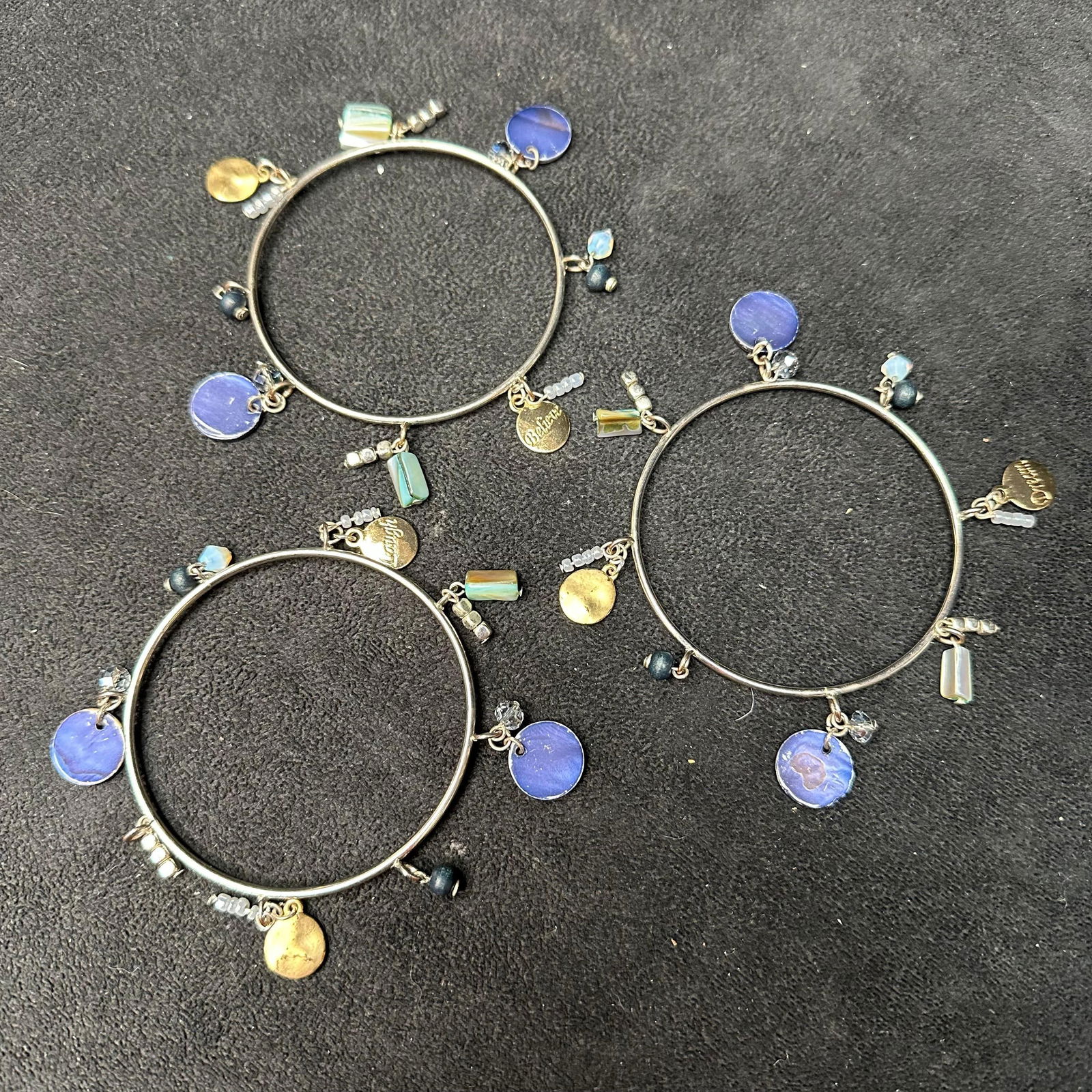 Set of Three Silver Tone Charm Bangle Bracelets with Blue and Gold Dangle Accents: Set of three coordinating silver-tone bangle bracelets featuring assorted dangling charms. Each bracelet is accented with blue enamel or glass discs, faceted beads, small metallic spacers, and