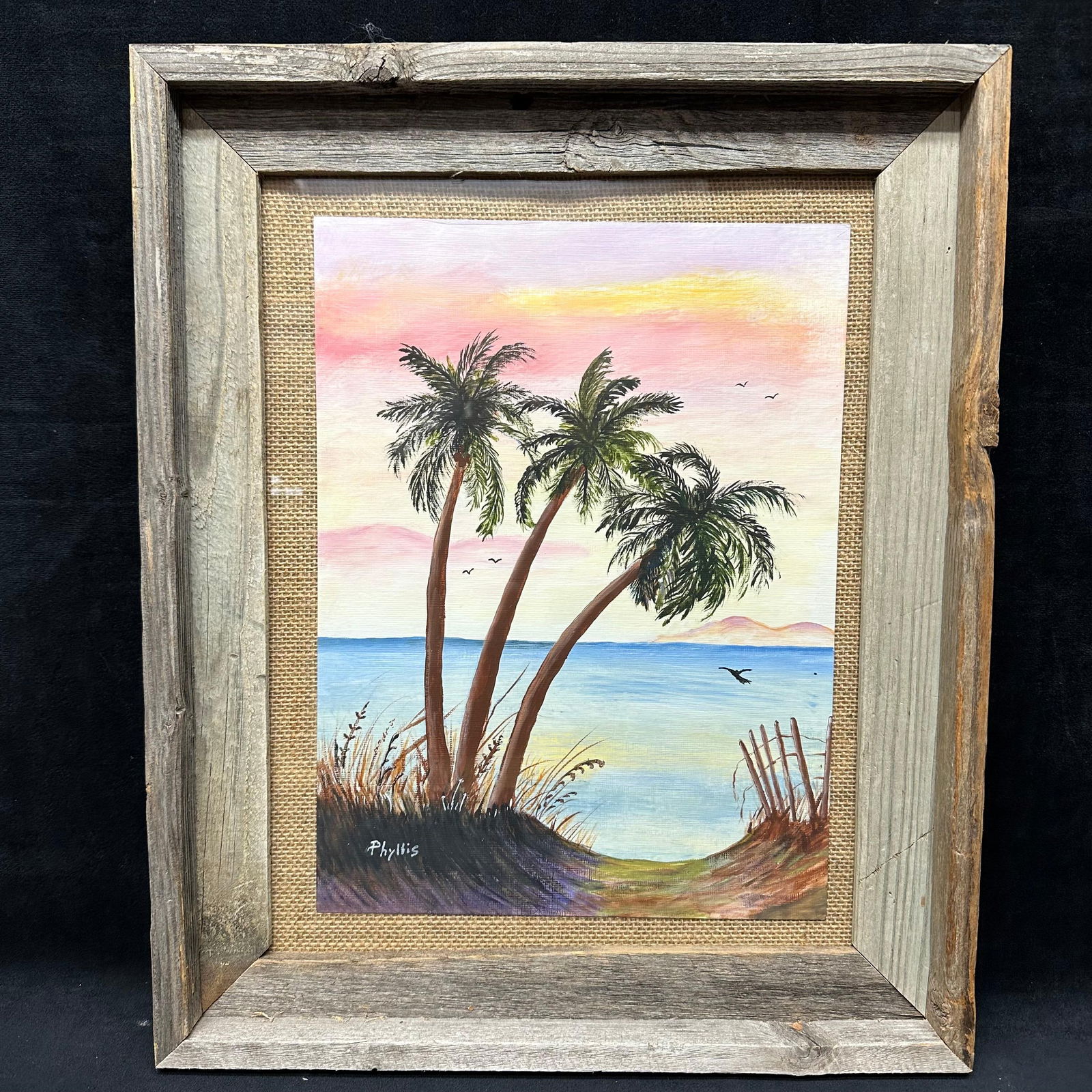Phyllis Signed Coastal Palm Tree Painting in Rustic Wood Frame: Original hand-painted coastal landscape signed by artist Phyllis, in the lower left. The scene depicts three palm trees overlooking water at sunset with birds in flight and dune fencing in the foregro