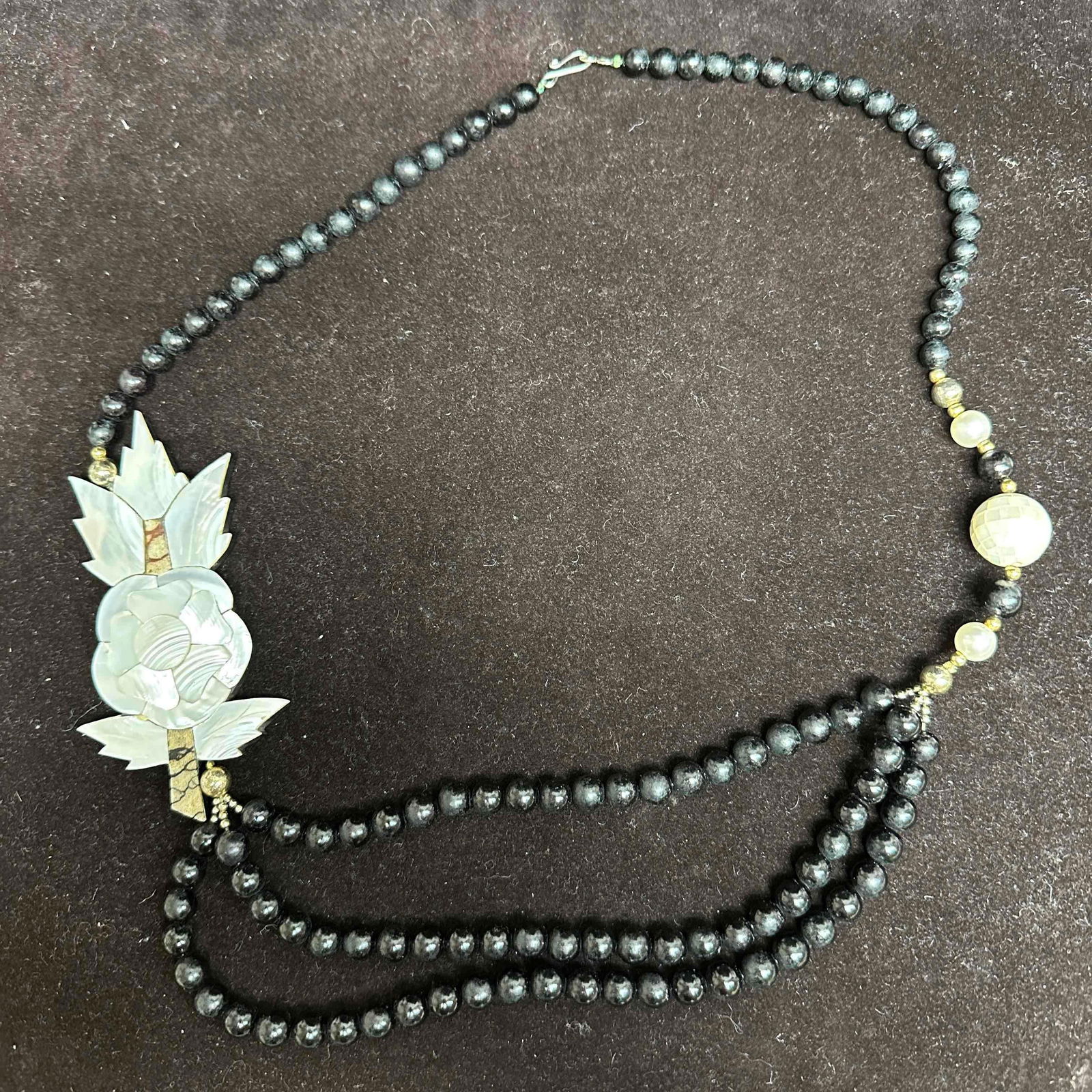 Mother Of Pearl Floral Pendant Necklace with Black Wooden Beaded Triple Strand: Necklace with black colored wooden beads with a beautiful mother of pearl pendant floral design. The necklace features floral and mother of pearl accents. The necklace features a j hook clasp