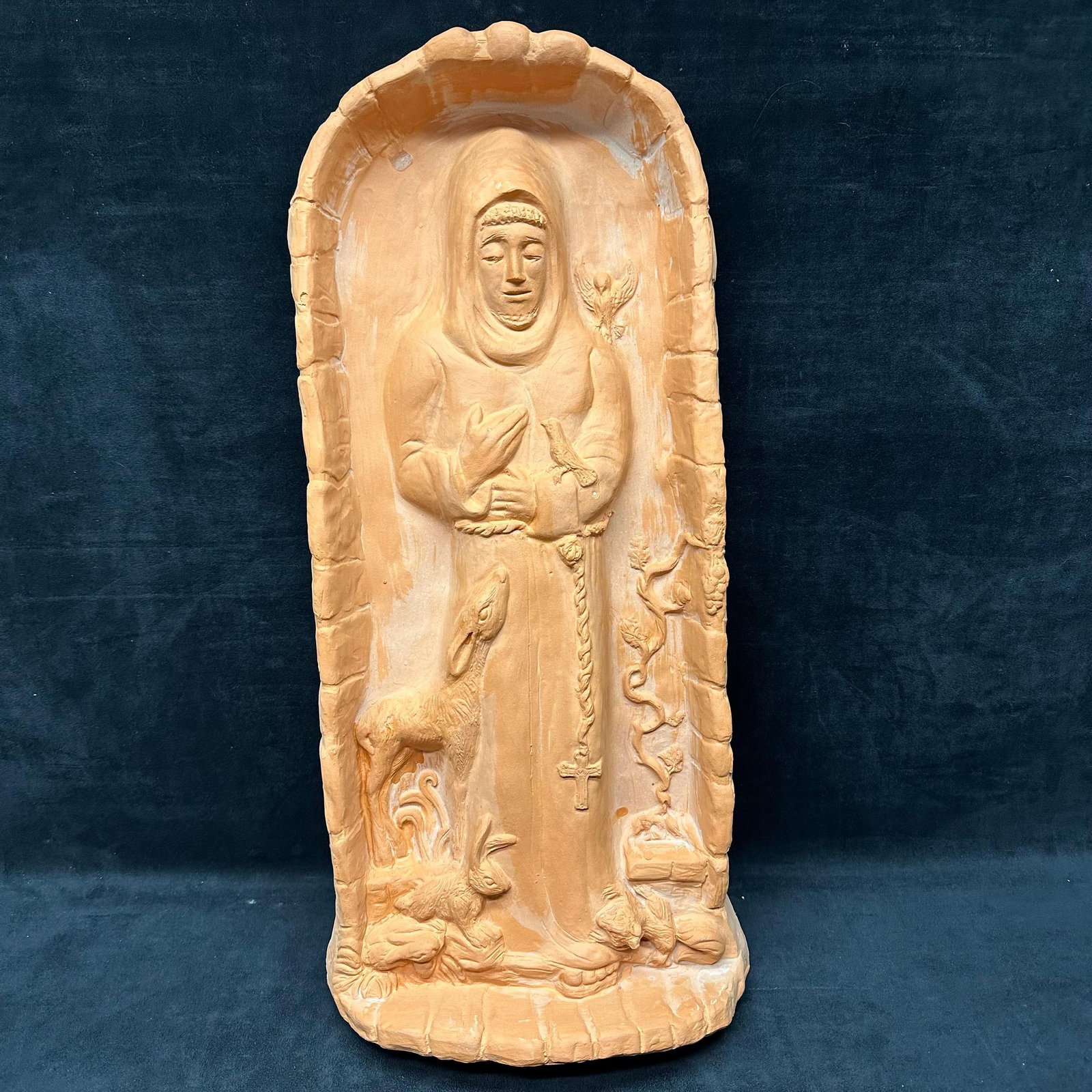 Terracotta Relief Plaque of Saint Francis: Medium terracotta wall plaque decor depicting Saint Francis of Assisi standing within an arched niche. Figure shown holding a rosary with a cross, surrounded by birds, a deer, and natural motifs in ra