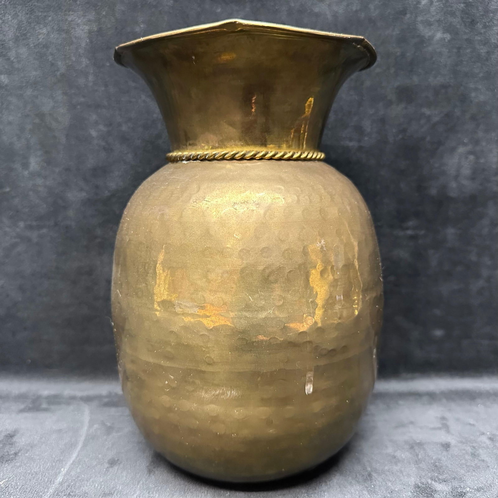 Medium Vintage Hand Hammered Brass Vase with Rope and Tassel Detail: Vintage brass vase featuring a hand-hammered body and wide flared rim. The design includes a cast rope-and-tassel relief wrapping around the shoulder. Displays surface oxidation, tarnish, and small de