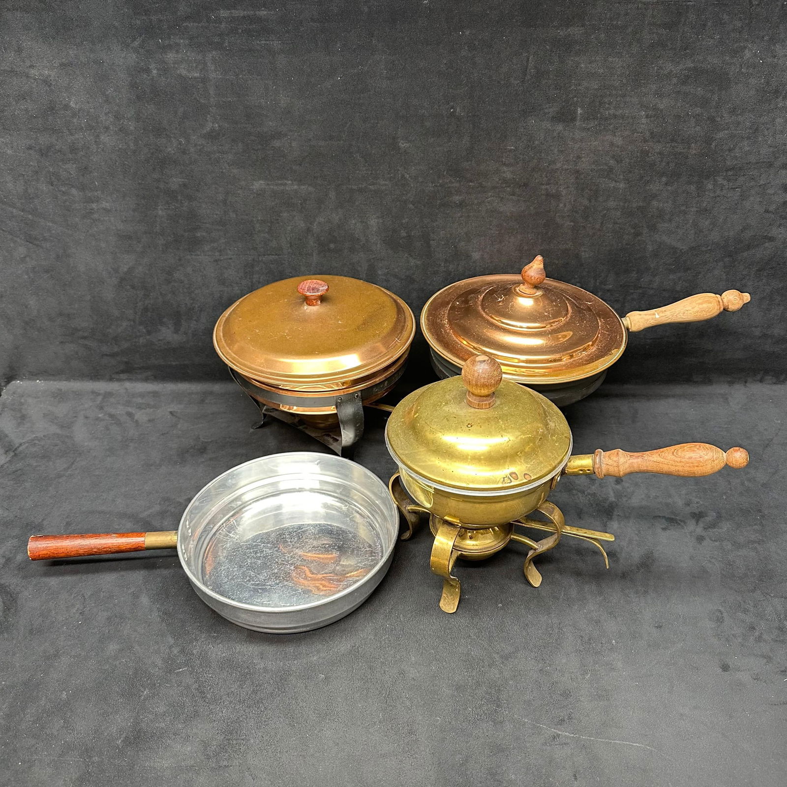 Vintage Brass Copper and Aluminum Chafing Dishes with Wooden Handles 4 Pieces: A lot of four assorted vintage chafing dishes and pans, including brass, copper, and aluminum examples. Each features a handled pot with a lid, and most include metal stands and burners. Designs vary