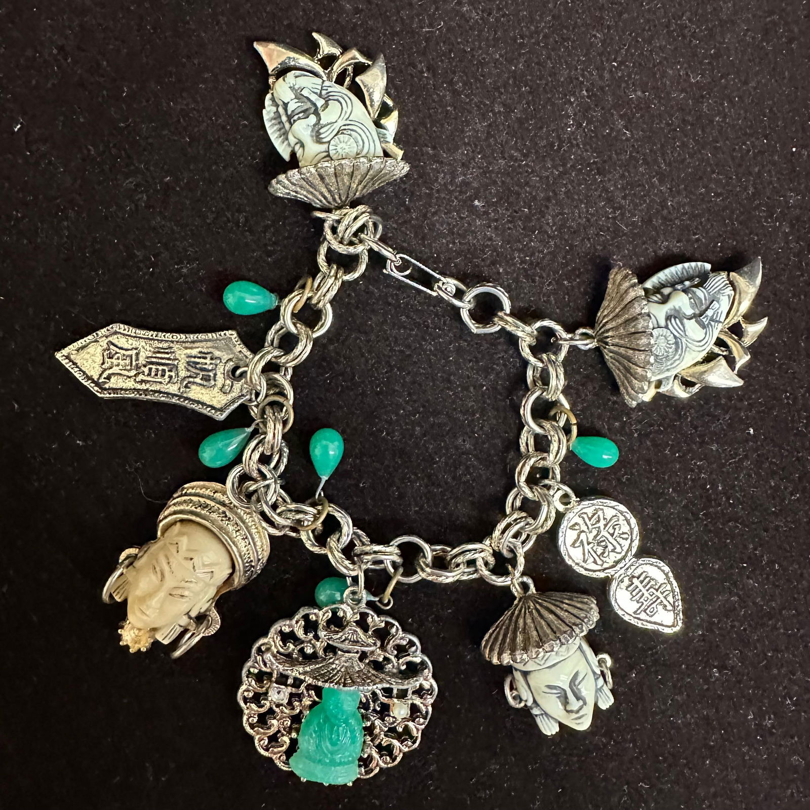 Asian Theme Charm Bracelet with Carved Figural Heads and Green Stone Drops: Vintage Asian inspired charm bracelet featuring multiple carved figural head charms, coin charms, character plaques, and green stone drops. The bracelet consists of a heavy brass/silver-tone base meta
