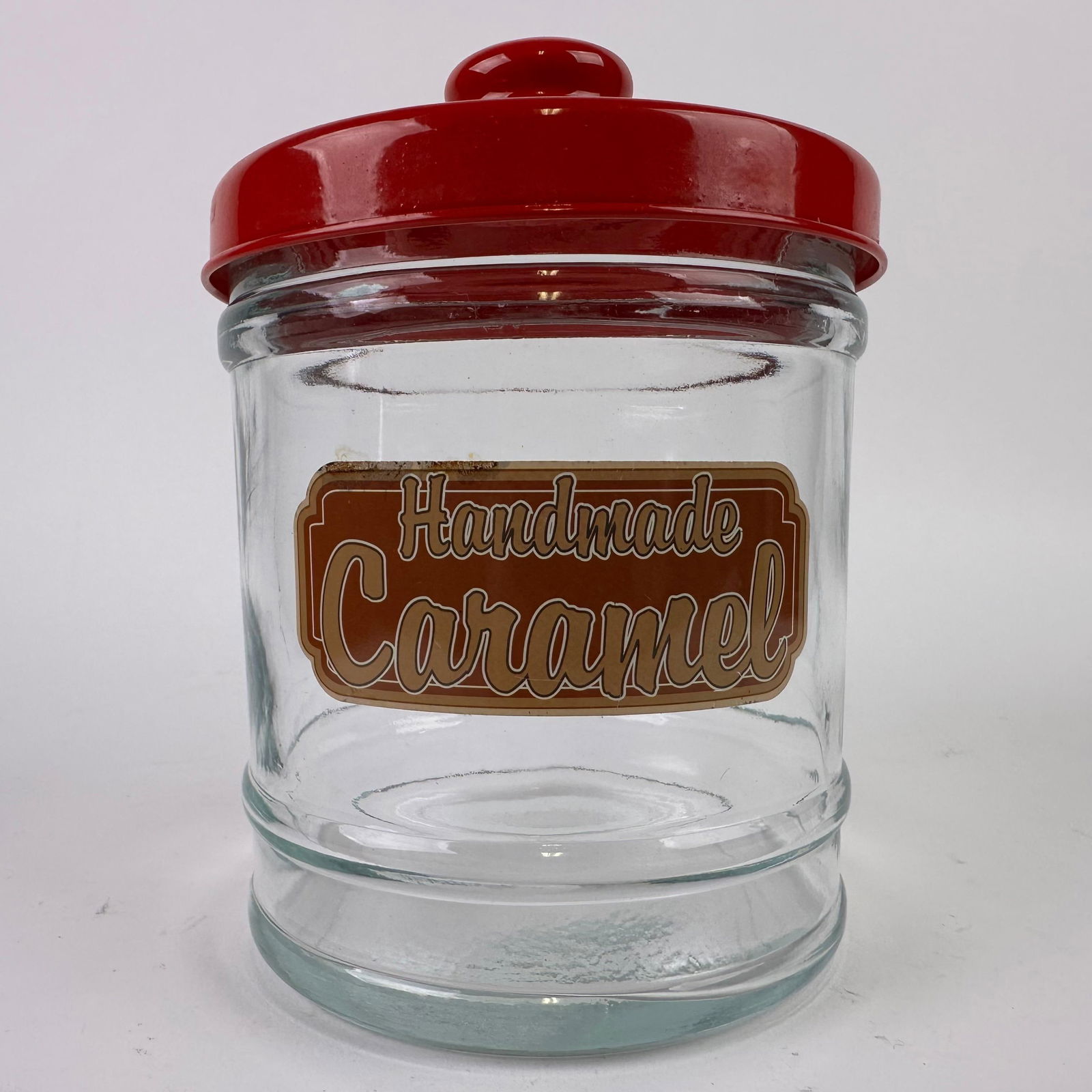 Vintage Style Glass Handmade Caramel Countertop Candy Jar with Red Lid: Vintage clear glass countertop candy jar with red plastic lid. The front retains the original label reading “Handmade Caramel.” Thick molded glass jar with stepped base and removable domed met