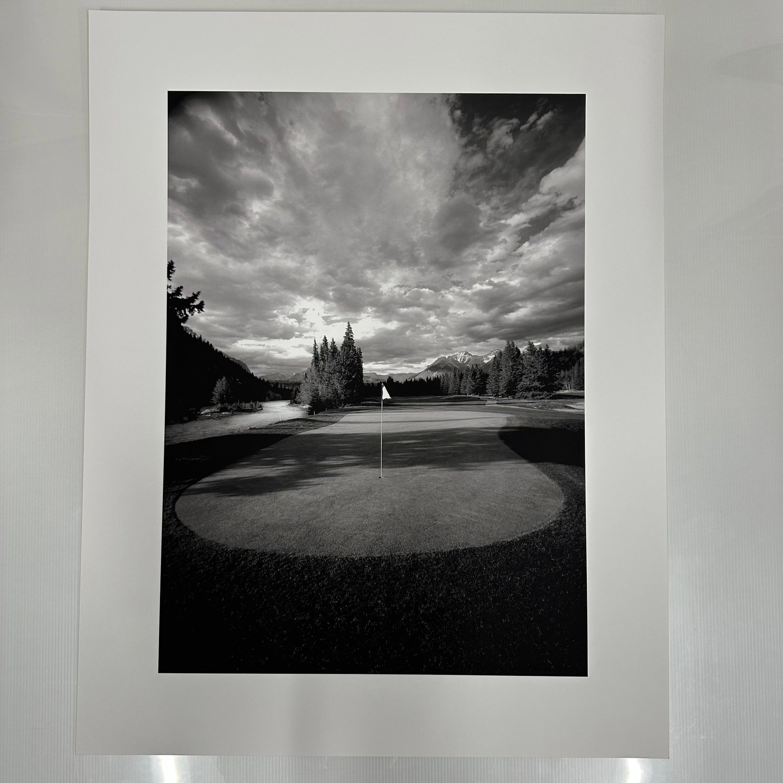 Banff Springs Golf Course in the Rocky Mountains of Canada Black and White Photo Print B: Large format black and white fine art photography print showing a golf green with a centered flag, surrounded by trees and distant mountains under a dramatic clouded sky. Printed on matte photo paper