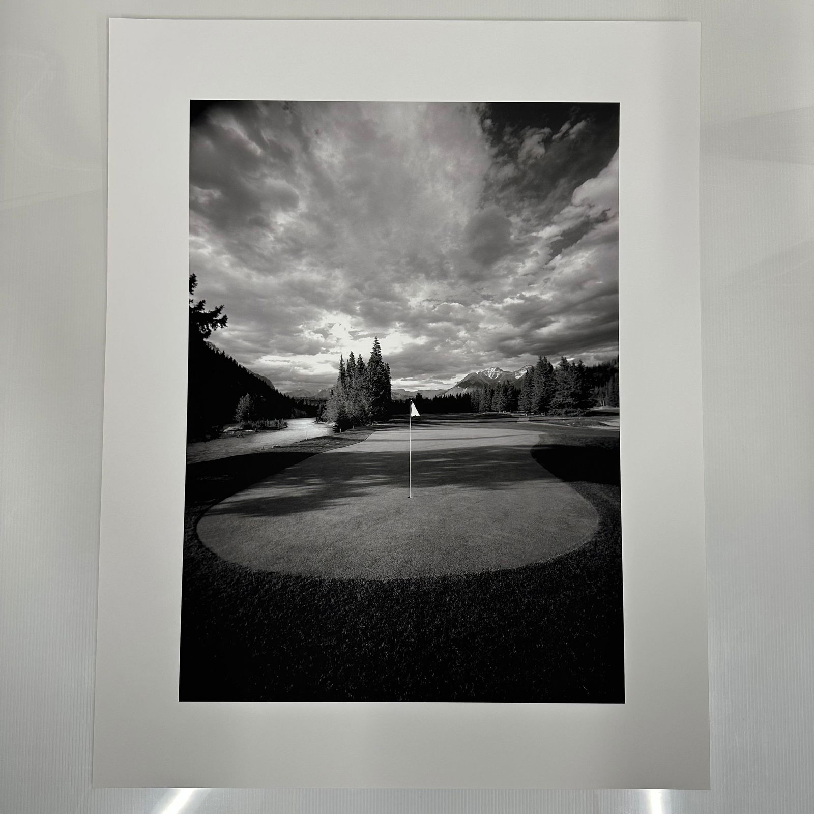 Banff Springs Golf Course in the Rocky Mountains of Canada Black and White Photo Print C: Large format black and white fine art photography print showing a golf green with a centered flag, surrounded by trees and distant mountains under a dramatic clouded sky. Printed on matte photo paper