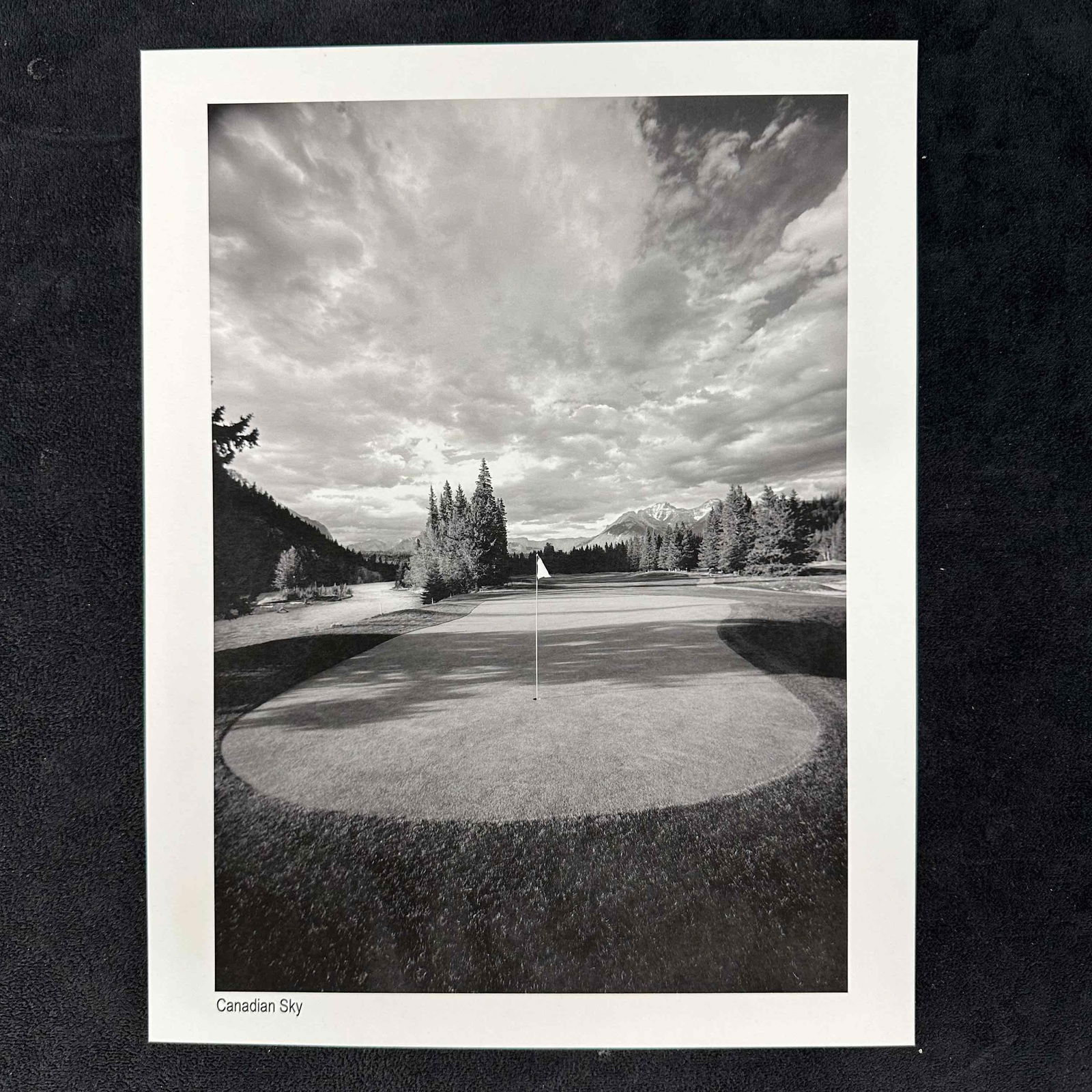 Canadian Sky Black and White Golf Photography Print: Black and white photograph titled Canadian Sky, featuring a beautifully framed golf green beneath a dramatic, cloud-filled sky with mountains in the distance. This item is in good pre-owned, previousl