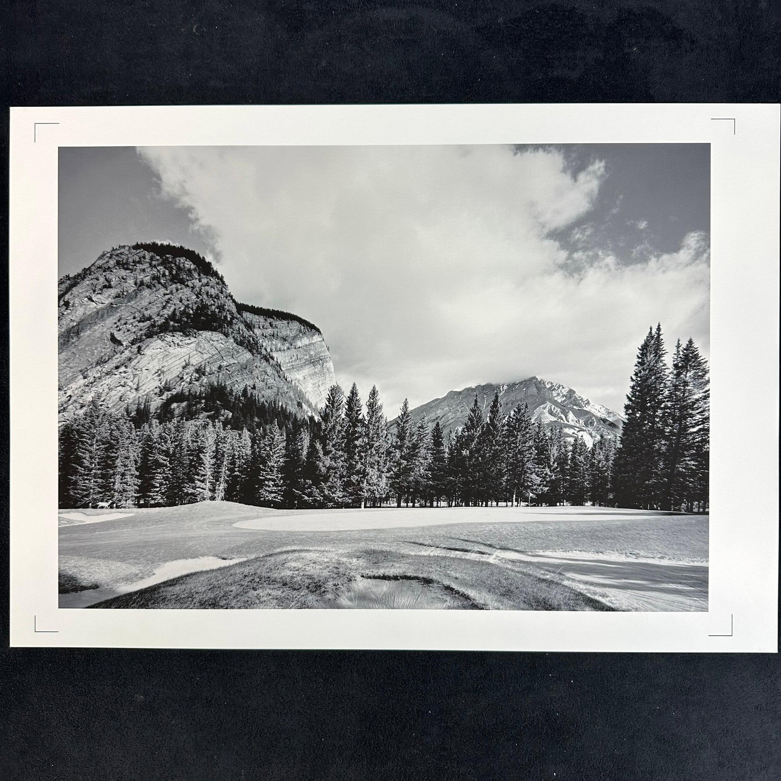 Mountain Golf Course Landscape Black and White Photography Print: Black and white landscape photography print capturing a golf course framed by towering mountains and dense evergreen forests under a partially clouded sky. Printed on high-quality photo paper, blank r
