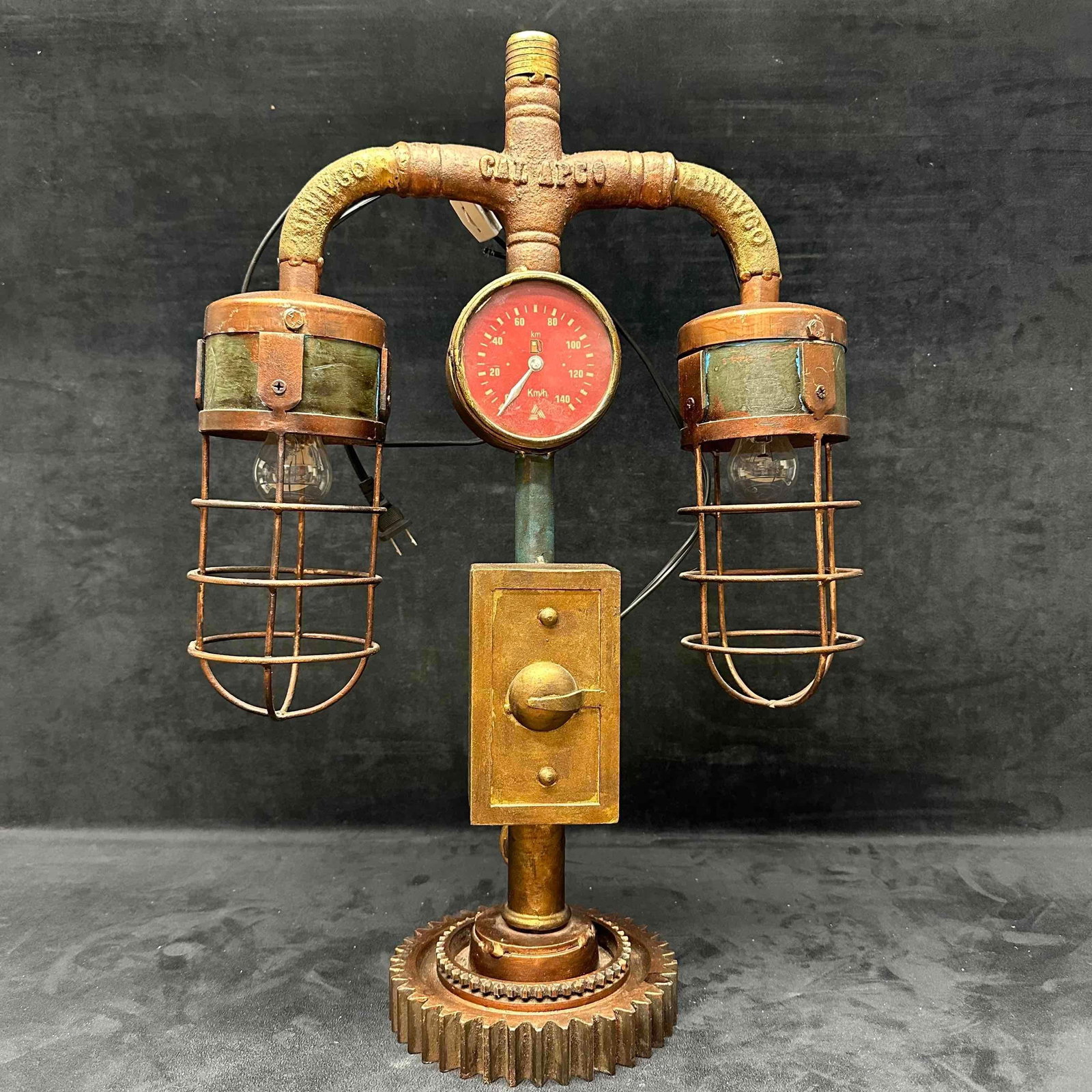 Industrial Steampunk Dual Cage Lamp with Vintage Gauge Piping and Gear Base: This unique industrial steampunk-style dual cage lamp crafted from repurposed metal components. Features two bulb sockets with protective wire cages, mounted on a curved pipe arm. At the center is a v