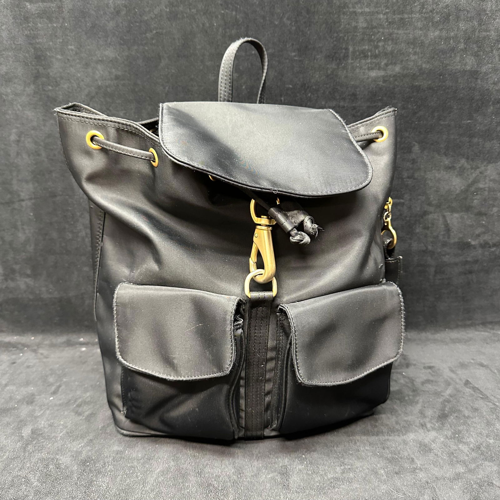 LJS Black Nylon Drawstring Backpack with Gold Tone Hardware: Black nylon backpack by LJS brand featuring a drawstring closure with flap top, gold-tone hardware, adjustable shoulder straps, dual exterior front pockets, and a zippered interior pocket. Interior