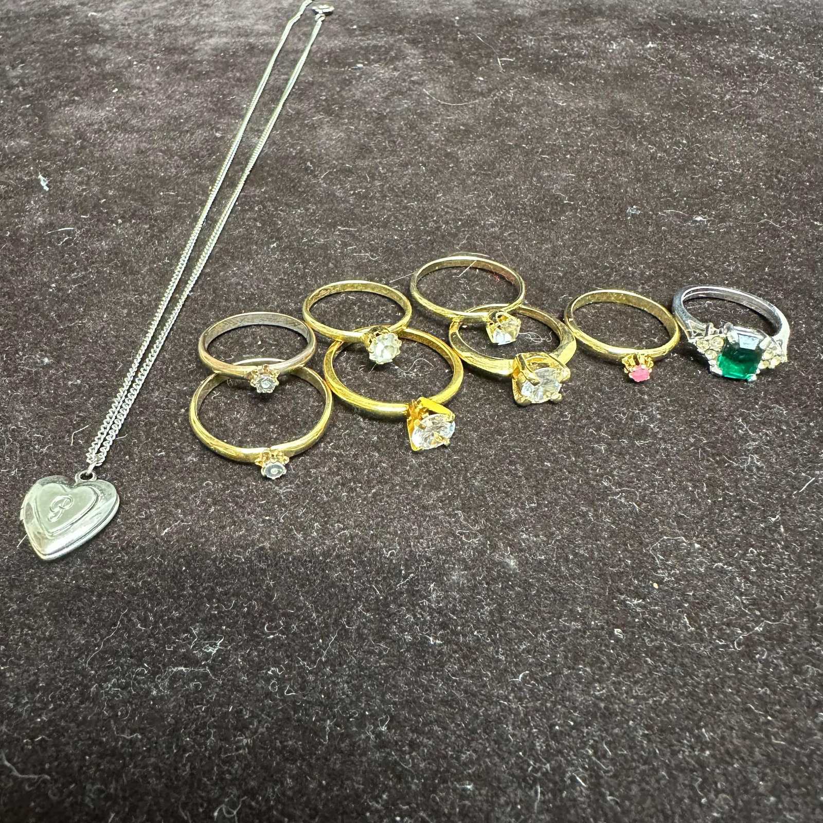 Collection of Costume Jewelry Rings and Silver Toned Heart Locket Necklace: An assorted group of costume jewelry, including seven gold-tone rings and one silver-tone ring set with simulated gemstones, featuring clear, pink, and green stones in various cuts. Also included is a