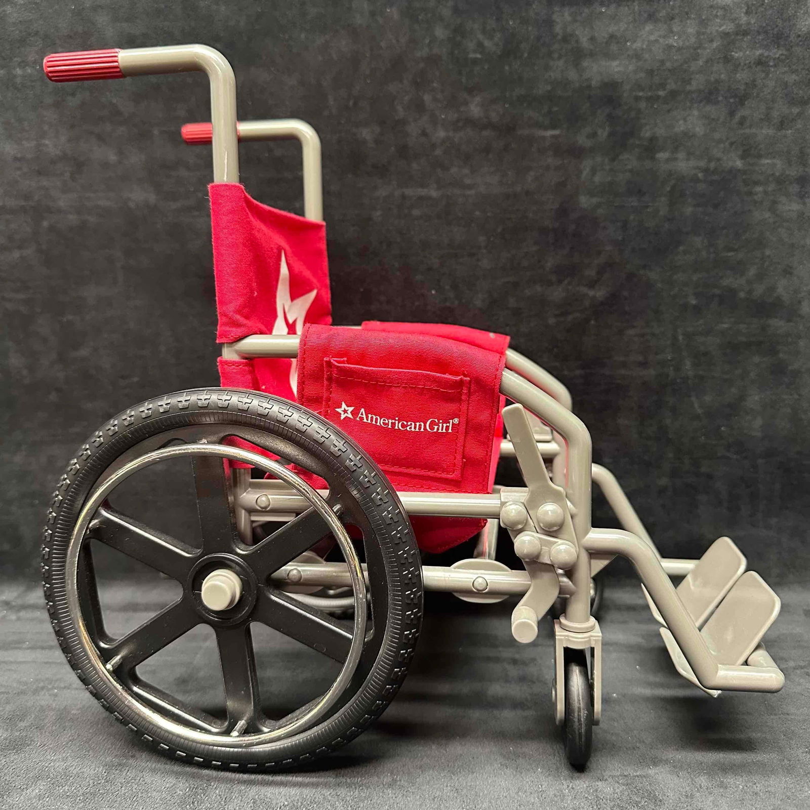 American Girl Doll Red Wheelchair Accessory: American Girl doll-sized wheelchair in gray with bright red fabric seat and backrest featuring the American Girl star logo. Functional design with large black wheels, movable footrests, and sturdy con