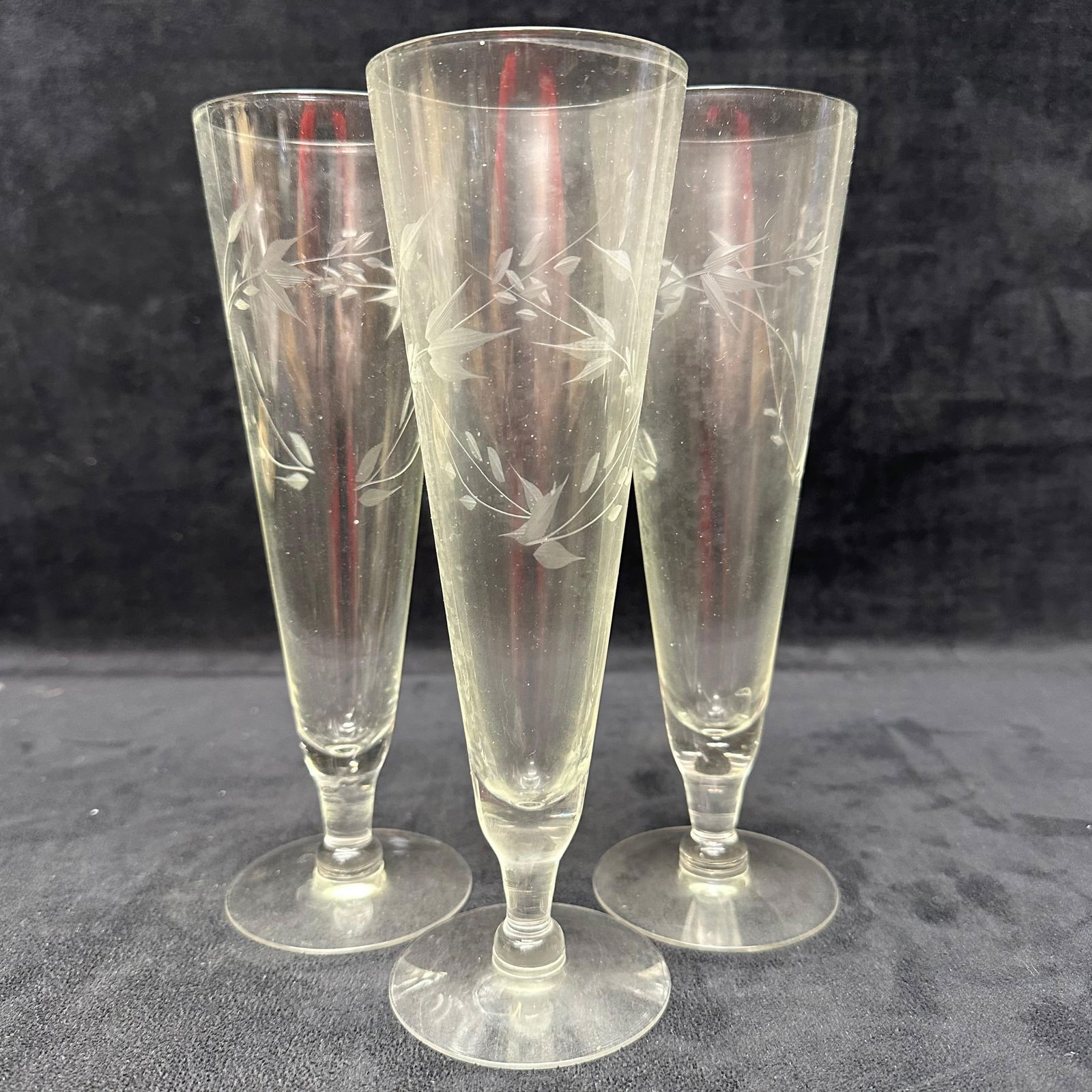 Set of Three Etched Glass Champagne Flutes with Floral Vine Motif Unmarked: Set of three clear glass champagne flutes featuring hand-etched floral and vine decoration around the bowls. Tall tapered forms with slender stems and round bases. Unmarked. These items are in good pr