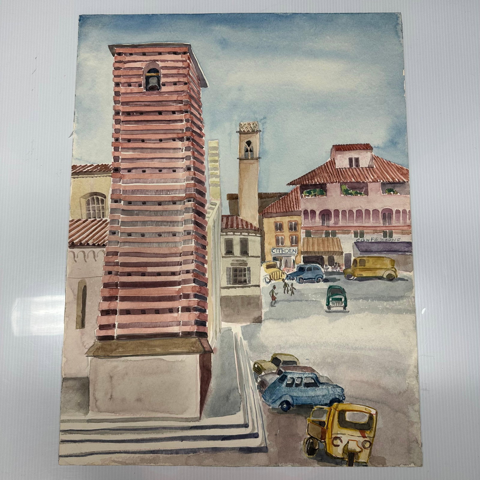 European Street Scene Watercolor Painting with Bell Tower and Vintage Automobiles: Unknown amateur artist Original watercolor painting depicting a European town square scene with striped brick bell tower, tiled roof buildings, storefronts, and vintage automobiles. No hallmark or sig