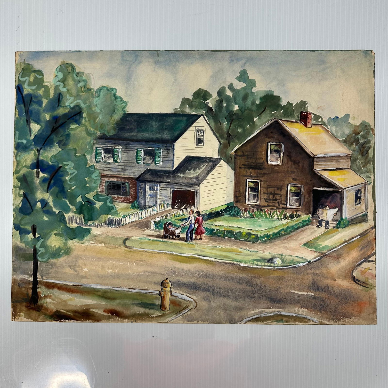 Original Watercolor Neighborhood Street Scene Inscribed Jo Swanson: Original watercolor painting depicting a neighborhood street scene with two houses, trees, figures walking with a baby carriage, and a fire hydrant at the corner. Painted on art paper. The reverse is