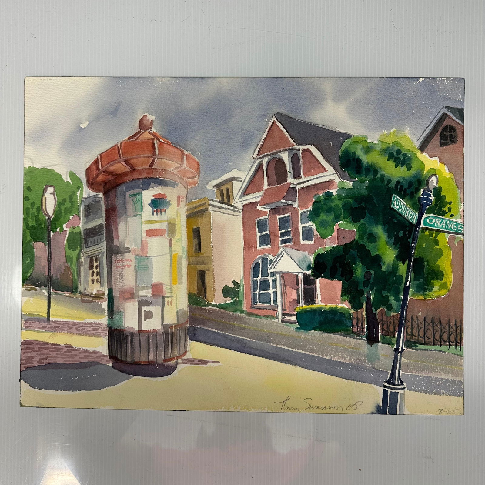 Thomas Swanson Street Scene Watercolor Painting Signed 1985: Original watercolor painting on paper depicting an urban street scene with historic architecture, trees, and a cylindrical advertising kiosk in the foreground. A street sign reading, Audubon and