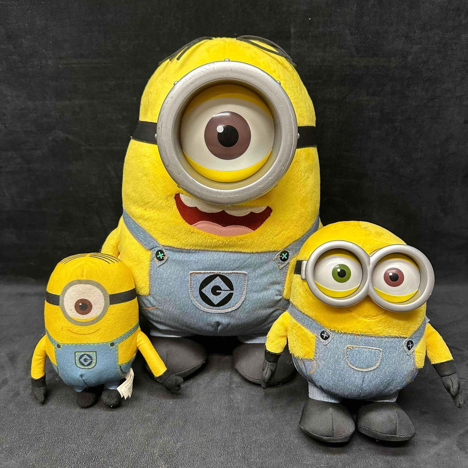 Lot of 3 Despicable Me Minions Plush Toys Assorted Sizes: Group of three Minions plush toys from the Despicable Me franchise. Includes two small soft plush characters and one large battery-operated plush with plastic eye/goggle detail and sound feature