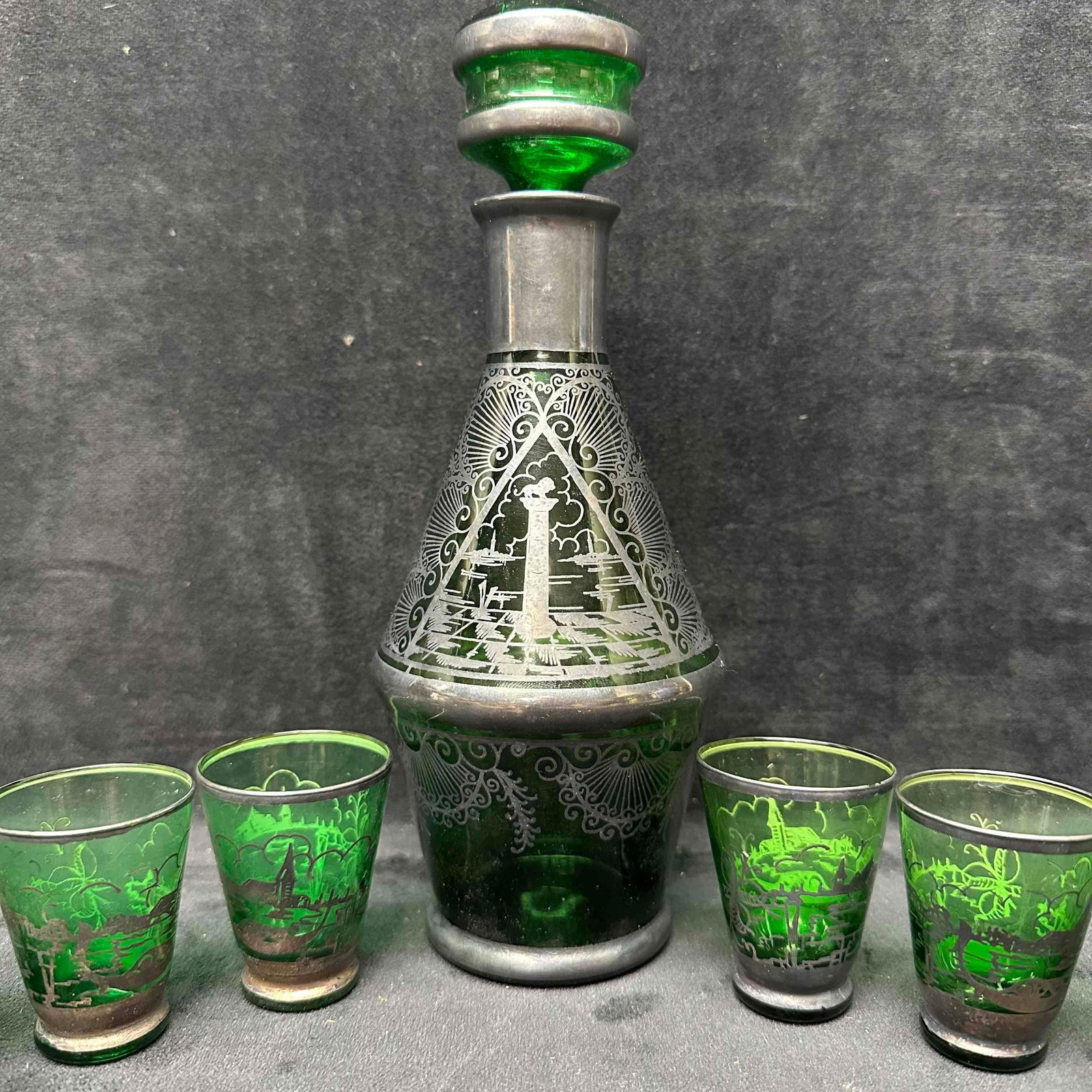 Green Glass Decanter with Six Matching Shot Glasses Silver Toned Etching Design: Vintage green glass decanter set with six matching shot glasses. The set features detailed silver toned overlay decoration with scenic designs. The decanter comes with its original stopper. All pieces