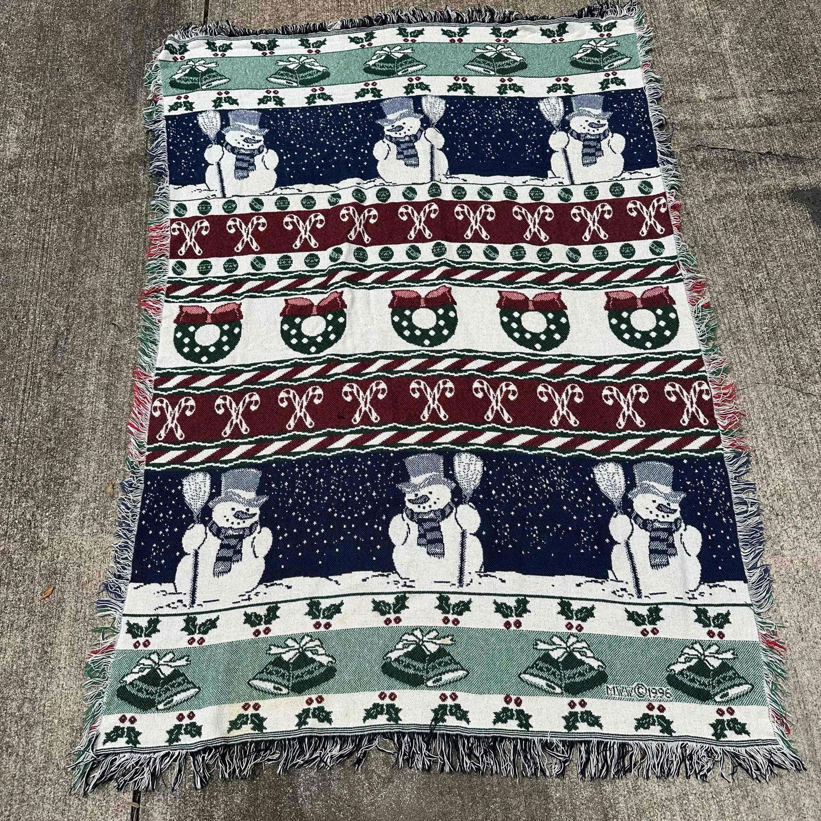 1996 Christmas Holiday Woven Throw Blanket with Snowman and Wreath Design: 1996 woven tapestry throw blanket featuring festive holiday motifs including snowmen, wreaths, candy canes, bells, and holly. The blanket has a colorful red, green, and blue design with decorative fri
