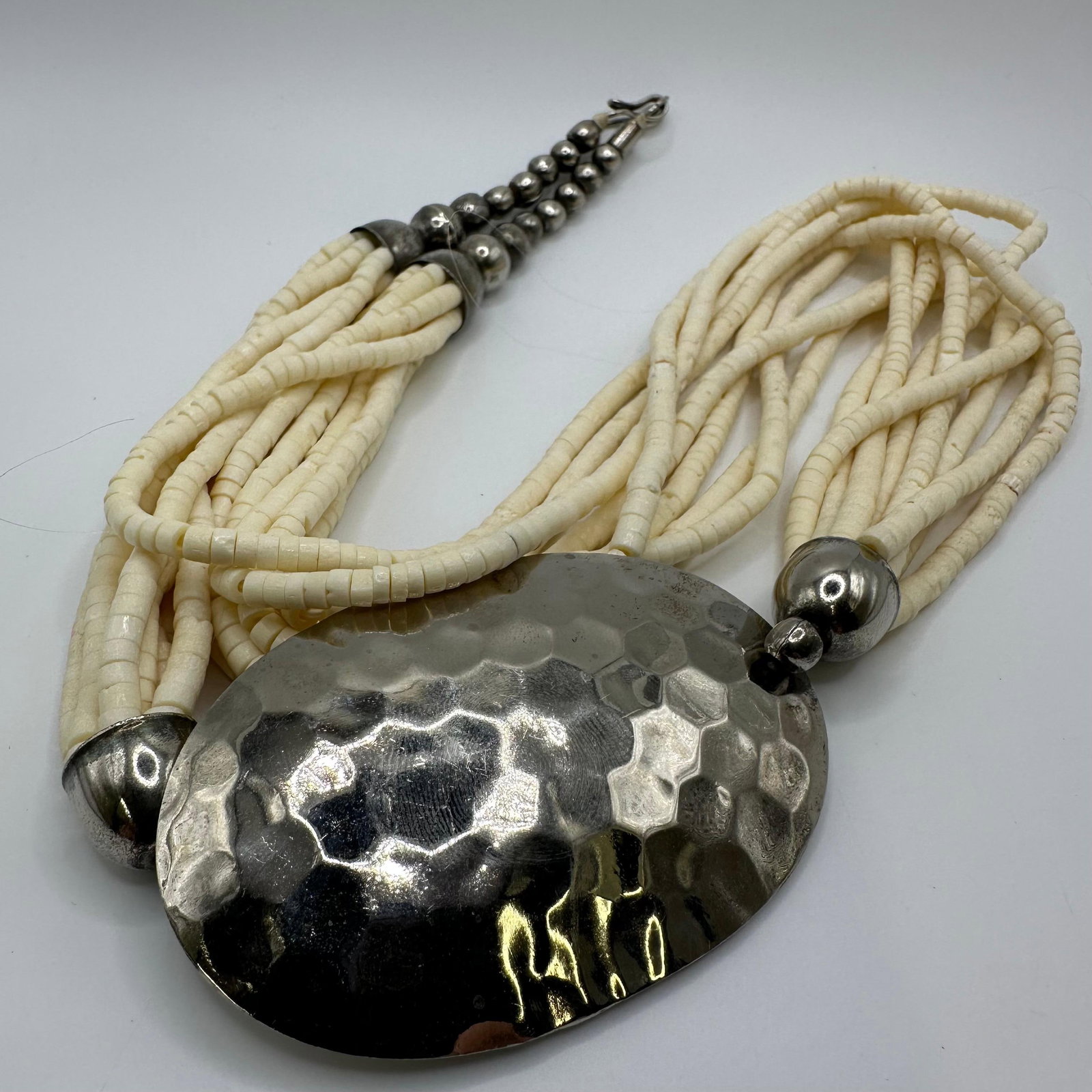 Ethnic African Tribal Style Bone Beaded Multi Strand Hammered Metal Accent Necklace: Statement style multi-strand necklace featuring multiple strands of bone beads, silver-tone hammered metal beads, and accents. Necklace includes approximately 8 strands of beading with alternating wh