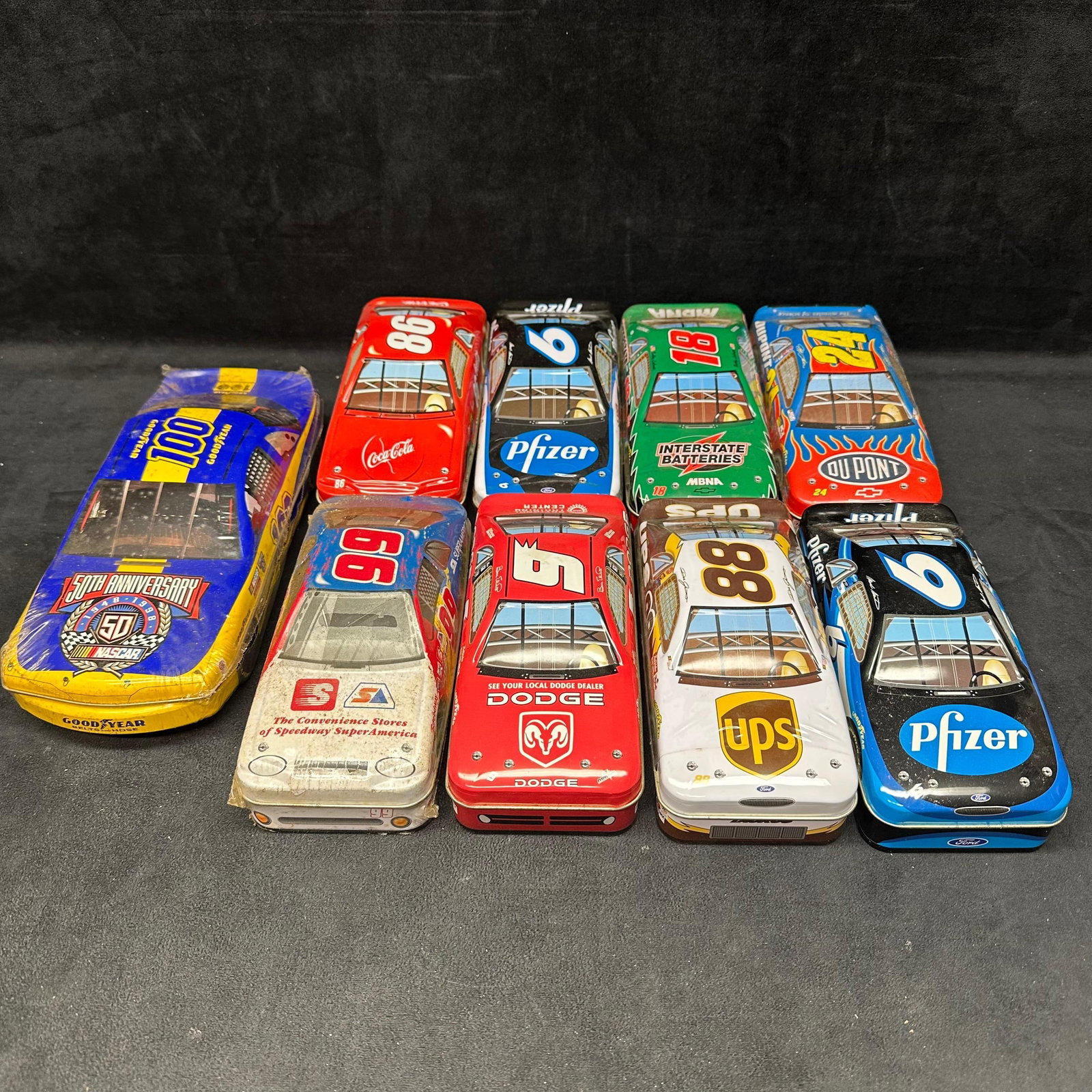 Lot of NASCAR Diecast Tin Cars Assorted Sponsors and Drivers: Collection of NASCAR diecast-style tin car containers featuring various sponsor designs, including DuPont, UPS, Pfizer, Interstate Batteries, Dodge, Coca-Cola, and others. Includes a NASCAR 50th Anniv
