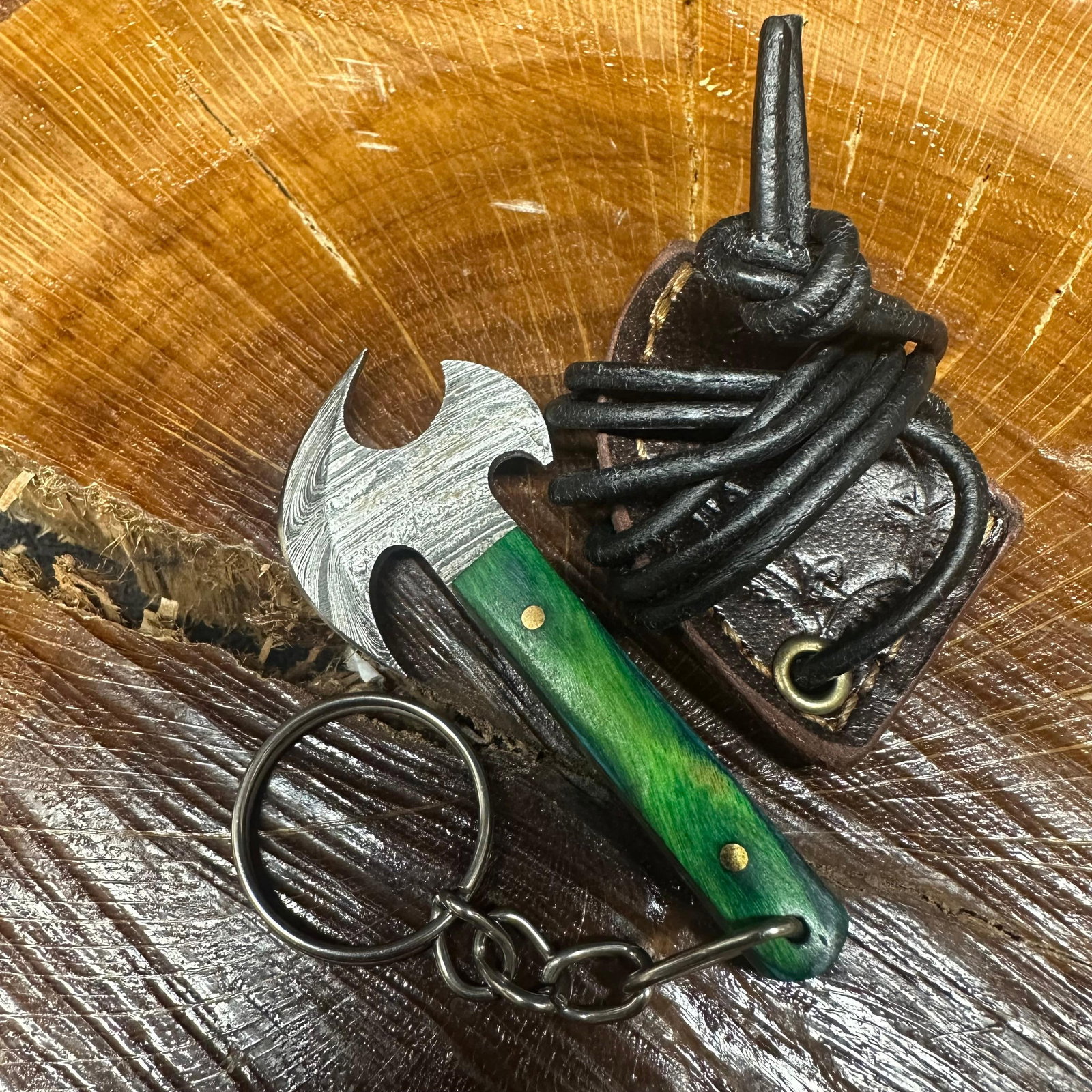 MHS Cutlery Keychain Layered Style Blade Axe With Leather Case Necklace R46: Small key chain-sized fixed-blade axe featuring a patterned steel blade and a compact epoxy-resin/wood green/blue-tone handle secured with brass pins. Includes a fitted brown leather sheath with stitc