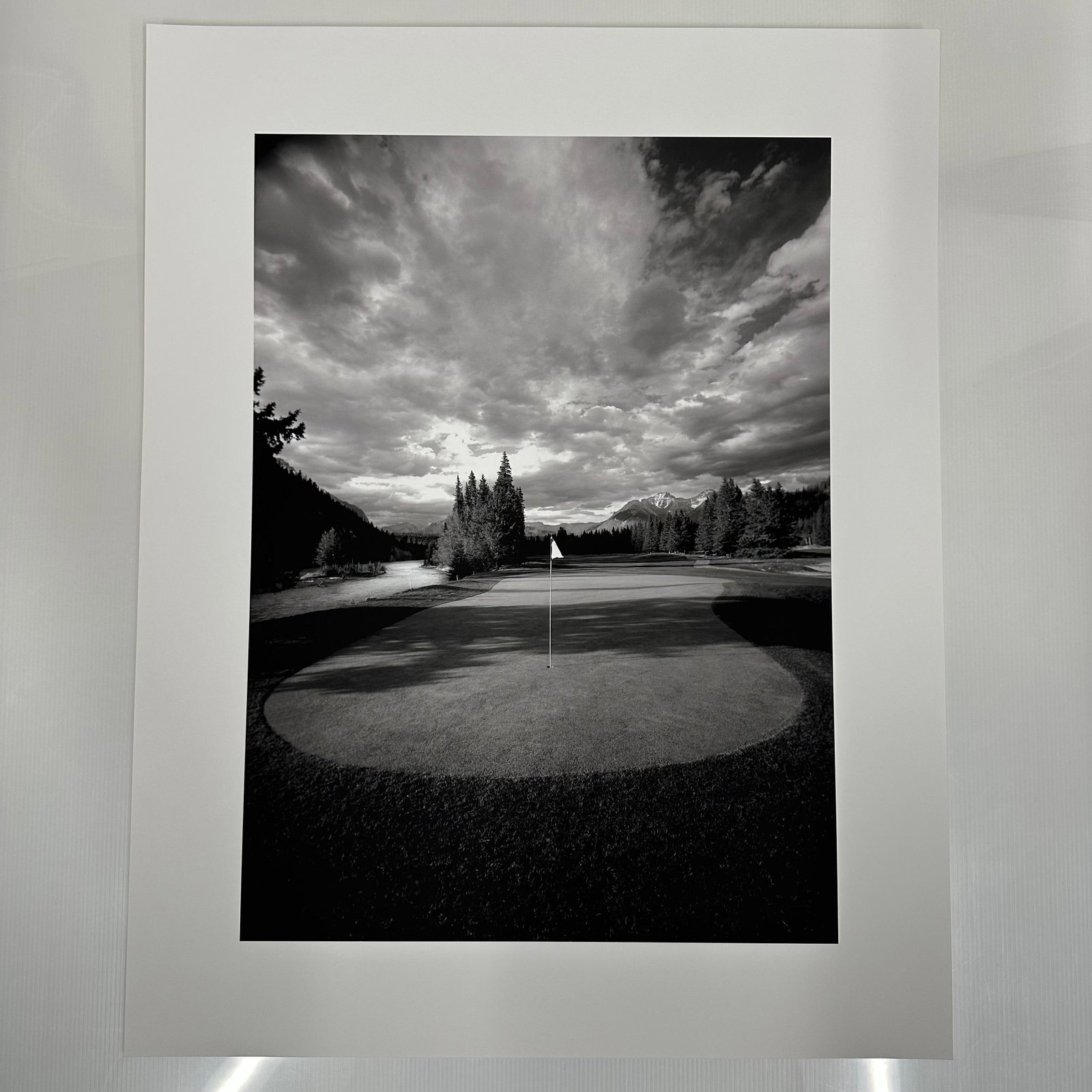 Banff Springs Golf Course in the Rocky Mountains of Canada Black and White Photo Print C: Large format black and white fine art photography print showing a golf green with a centered flag, surrounded by trees and distant mountains under a dramatic clouded sky. Printed on matte photo paper