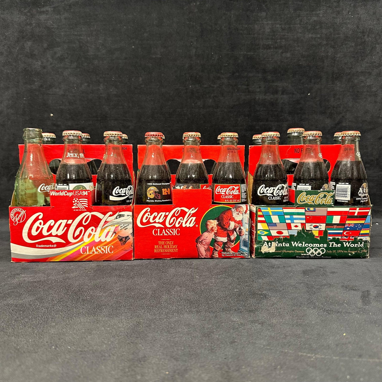 Coca Cola Commemorative 6 Pack Bottles with Carriers: Group of vintage Coca-Cola glass bottles in original cardboard 6-pack carriers. Includes multiple commemorative themes such as the Atlanta Olympics, the World Cup USA '94, and holiday designs. Bot