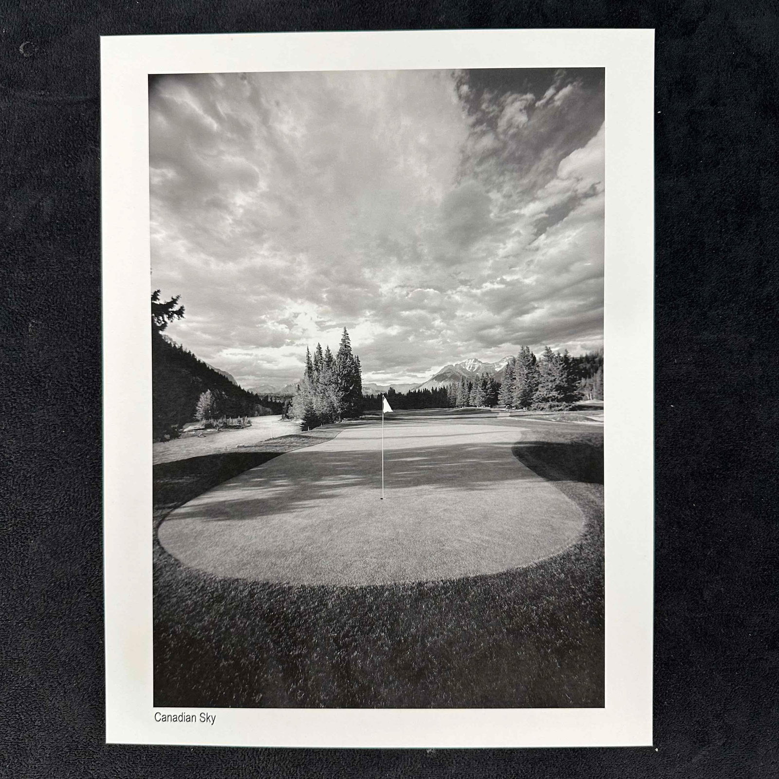 Canadian Sky Black and White Golf Photography Print: Black and white photograph titled Canadian Sky, featuring a beautifully framed golf green beneath a dramatic, cloud-filled sky with mountains in the distance. This item is in good pre-owned, previousl