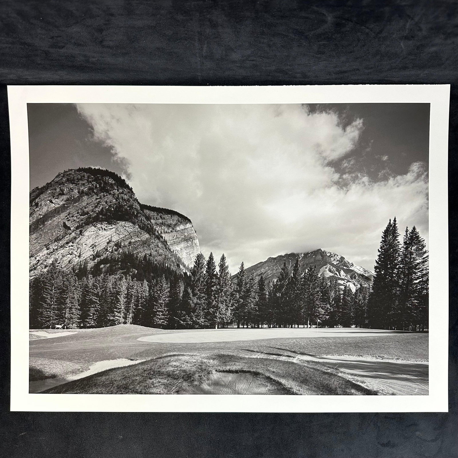 Black and White Inkjet Photograph Print Mountain Golf Course with Sheer Cliff Face: Black and white inkjet print depicting a golf course framed by towering cliffs and evergreen forest. The rugged rock face rises behind the tree line, evoking the grandeur of alpine landscapes. Wide to