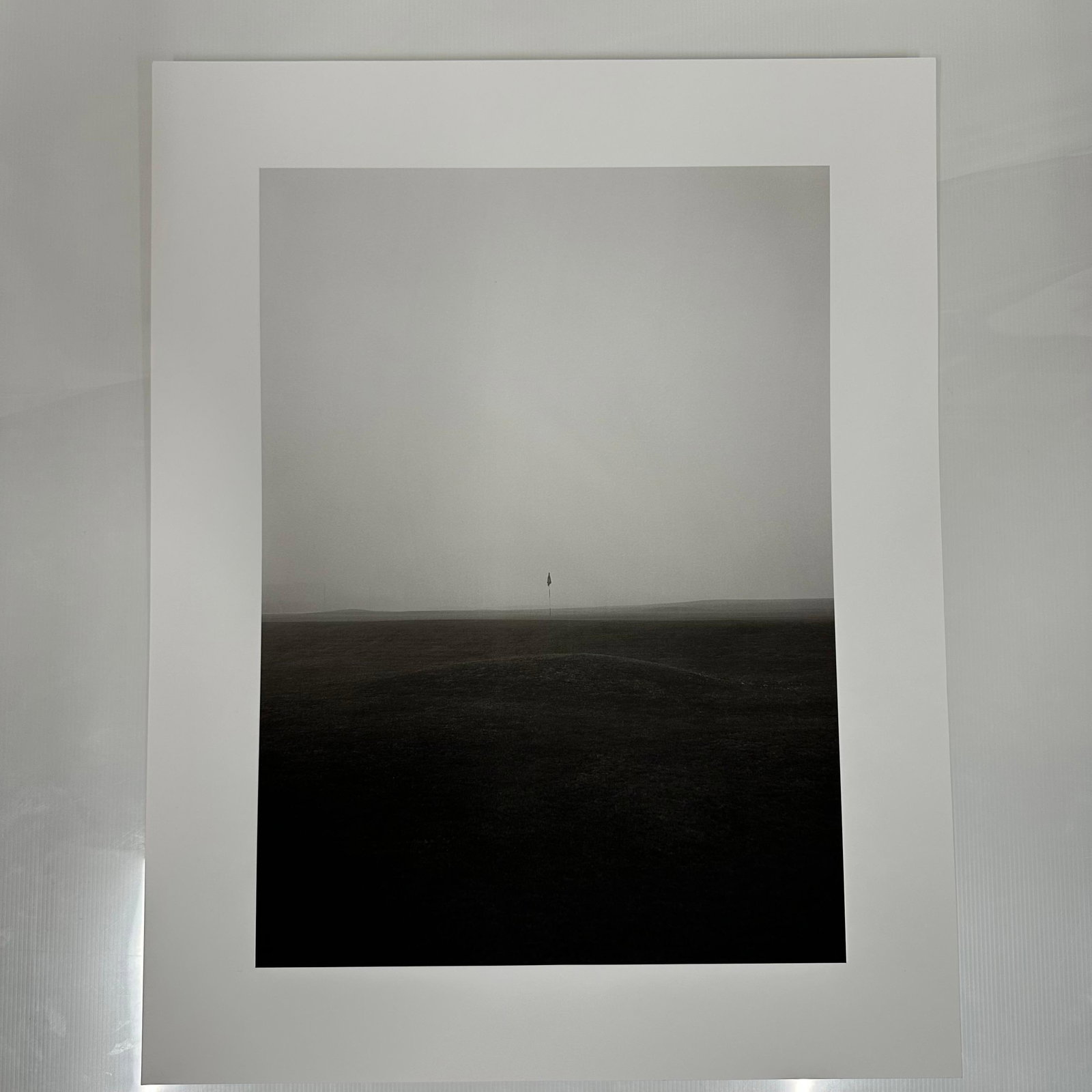 Foggy Golf Course Fairway Scene Black and White Minimalist Photography Print #1: Black and white photographic print depicting a solitary golf flag standing on a mist-covered fairway. The minimalist composition and muted tones create a serene, almost abstract landscape. Printed on