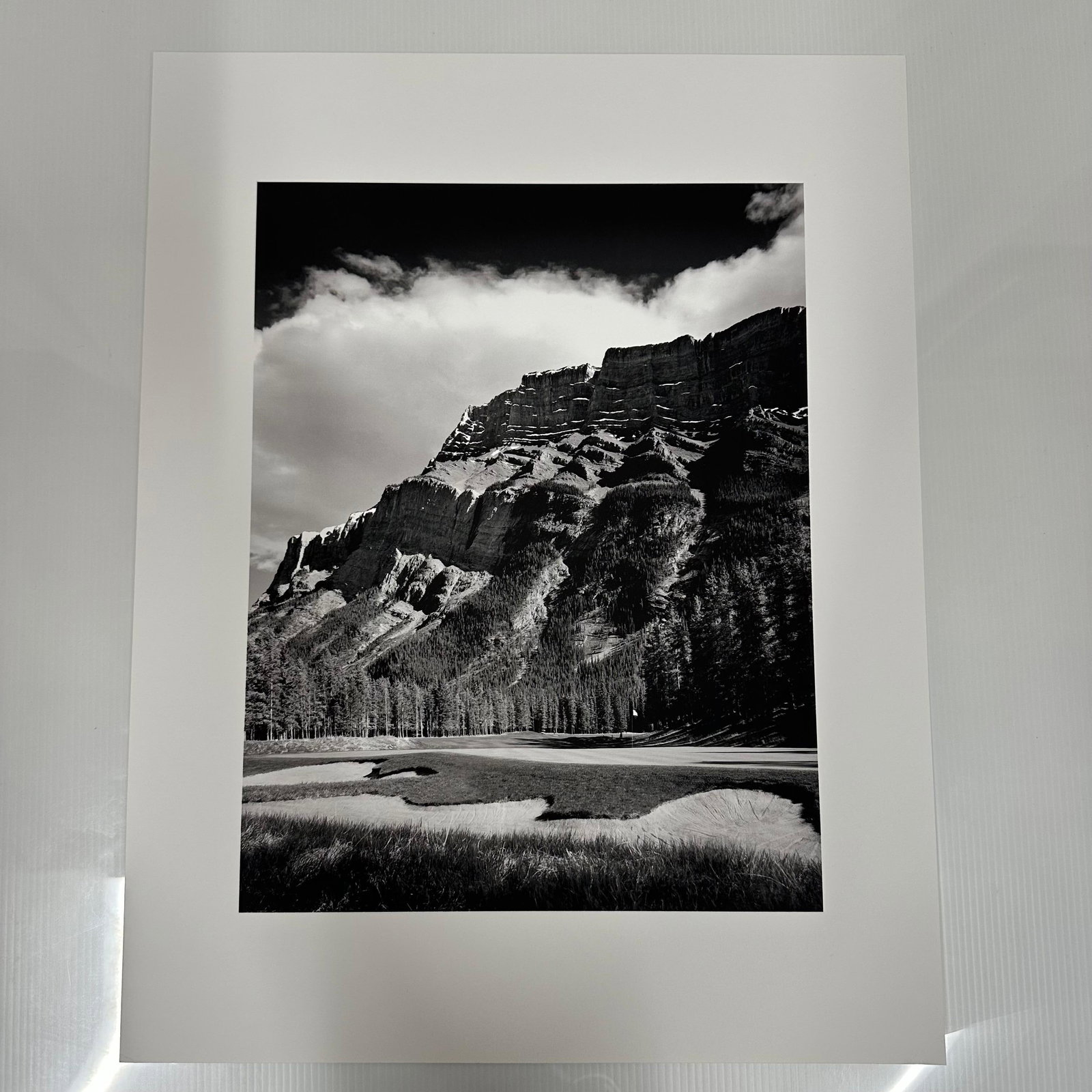 Black and White Landscape Photography Print Mountain Golf Course Scene: Black and white photographic print depicting a dramatic mountain range rising above a golf course landscape, possibly the Banff Springs Golf Course in Alberta, Canada. The image captures the contrast