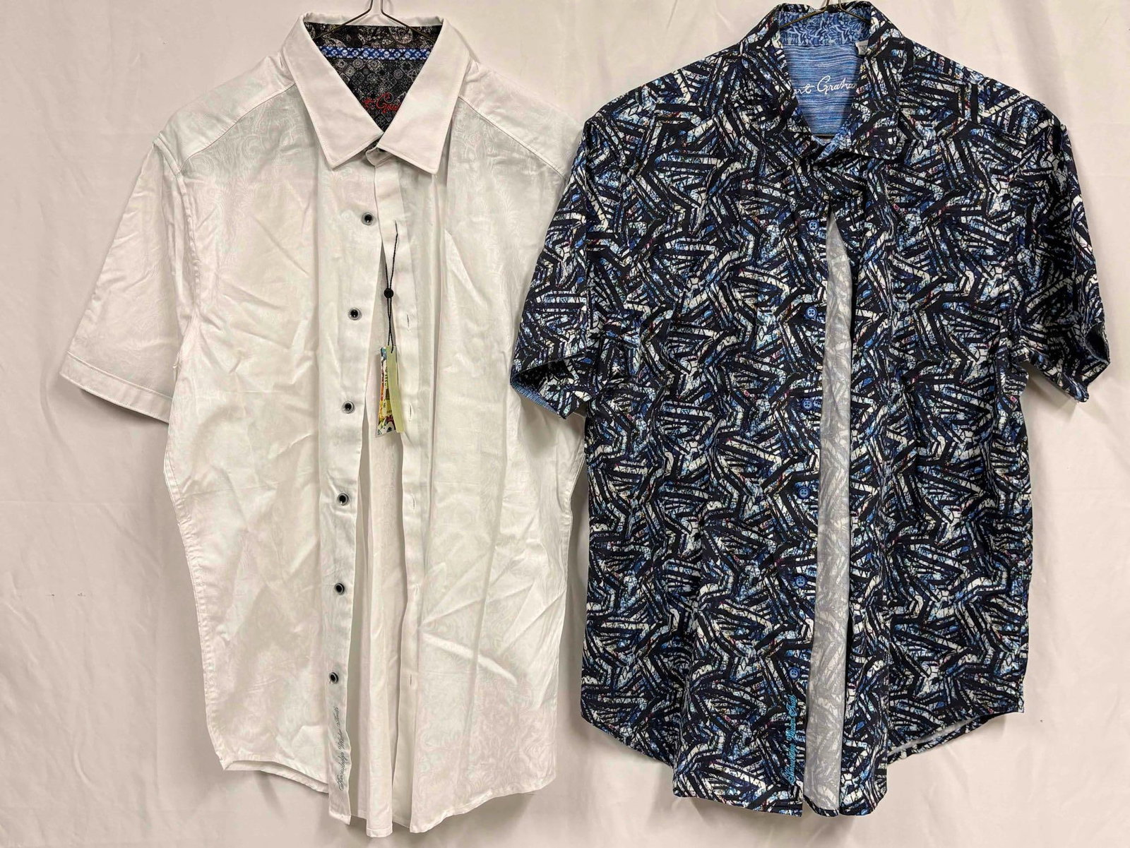 2PC Robert Grahams White and Blue Patterned Mens Short Sleeve Dress Shirt Bundle: This lot contains a pair of Robert Graham's shirts, a high-end clothing collection known for their men's polos and button-downs. Both shirts standing in like-new conditions, one of which still