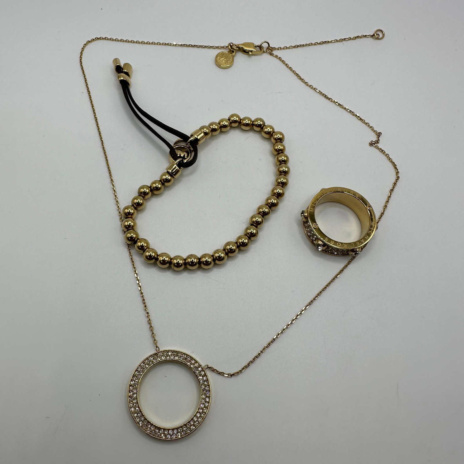 Michael Kors Gold Tone Jewelry Lot Necklace Bracelet And Ring Set: Group of Michael Kors gold tone fashion jewelry, including a pendant necklace with circular pave crystal accent, bead style bracelet with leather pull closure, an MK charm, and a matching pave crystal