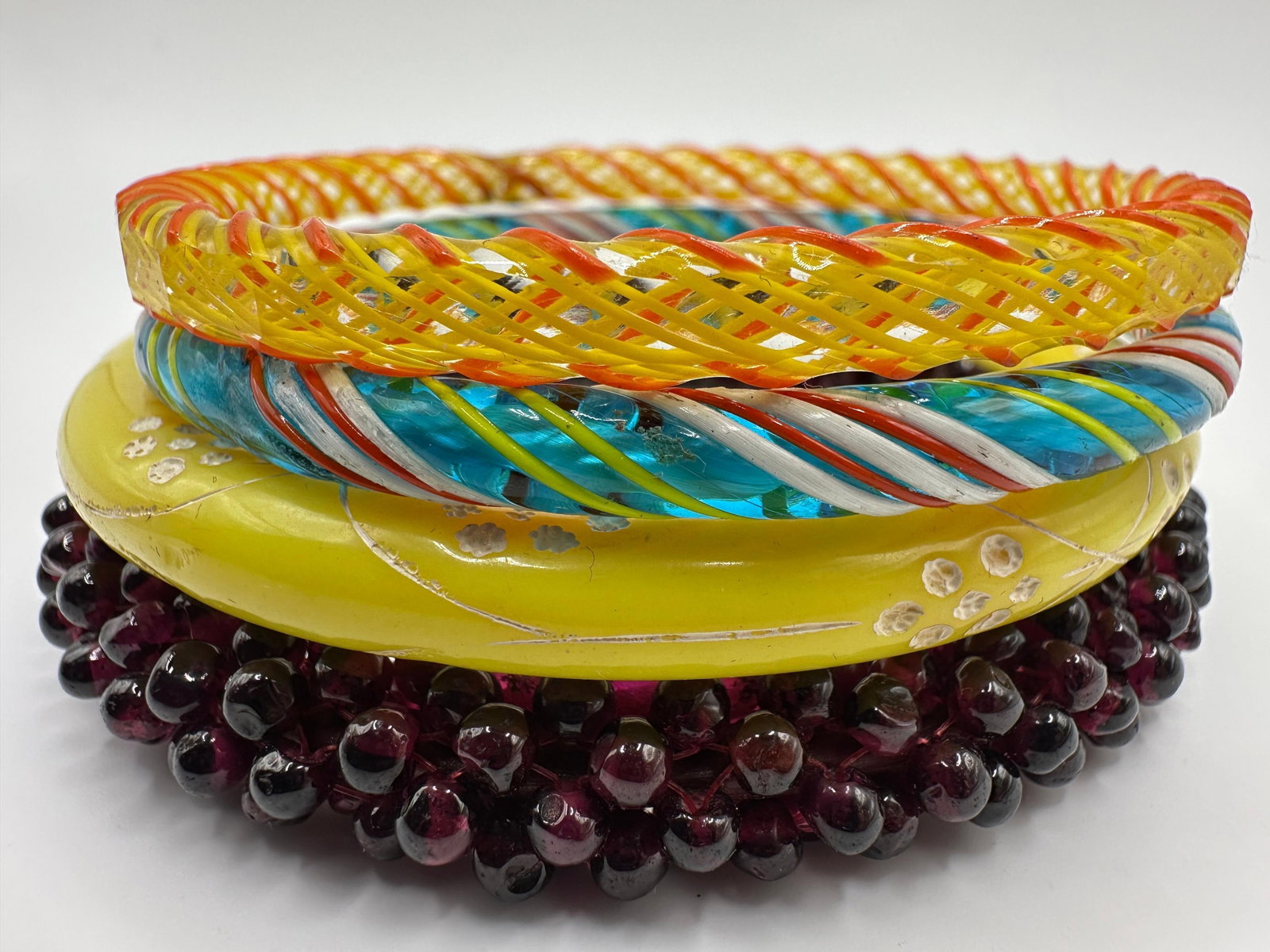 Lot of 4 Colorful Glass Bangles Latticino Style and Red Stone Beaded Bracelet: Group of four glass bangles, including a yellow example with applied white decoration, a multicolor twisted latticino style bangle, an openwork yellow and orange lattice bangle, and a dark red stone b