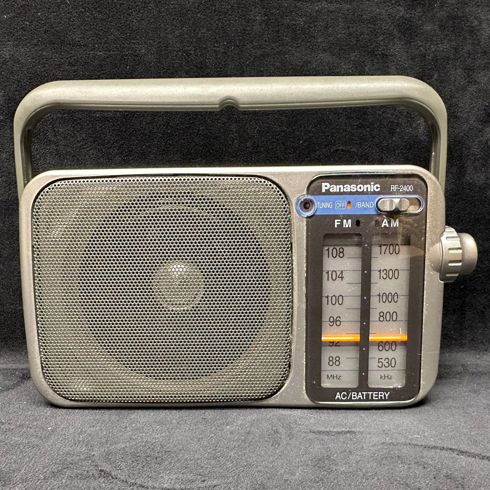 Panasonic RF-2400 Portable AM/FM Radio with Handle: Compact Panasonic RF-2400 portable radio featuring AM/FM reception with an analog tuning dial. Gray plastic housing with integrated carry handle, front speaker grille, and side tuning/volume controls.