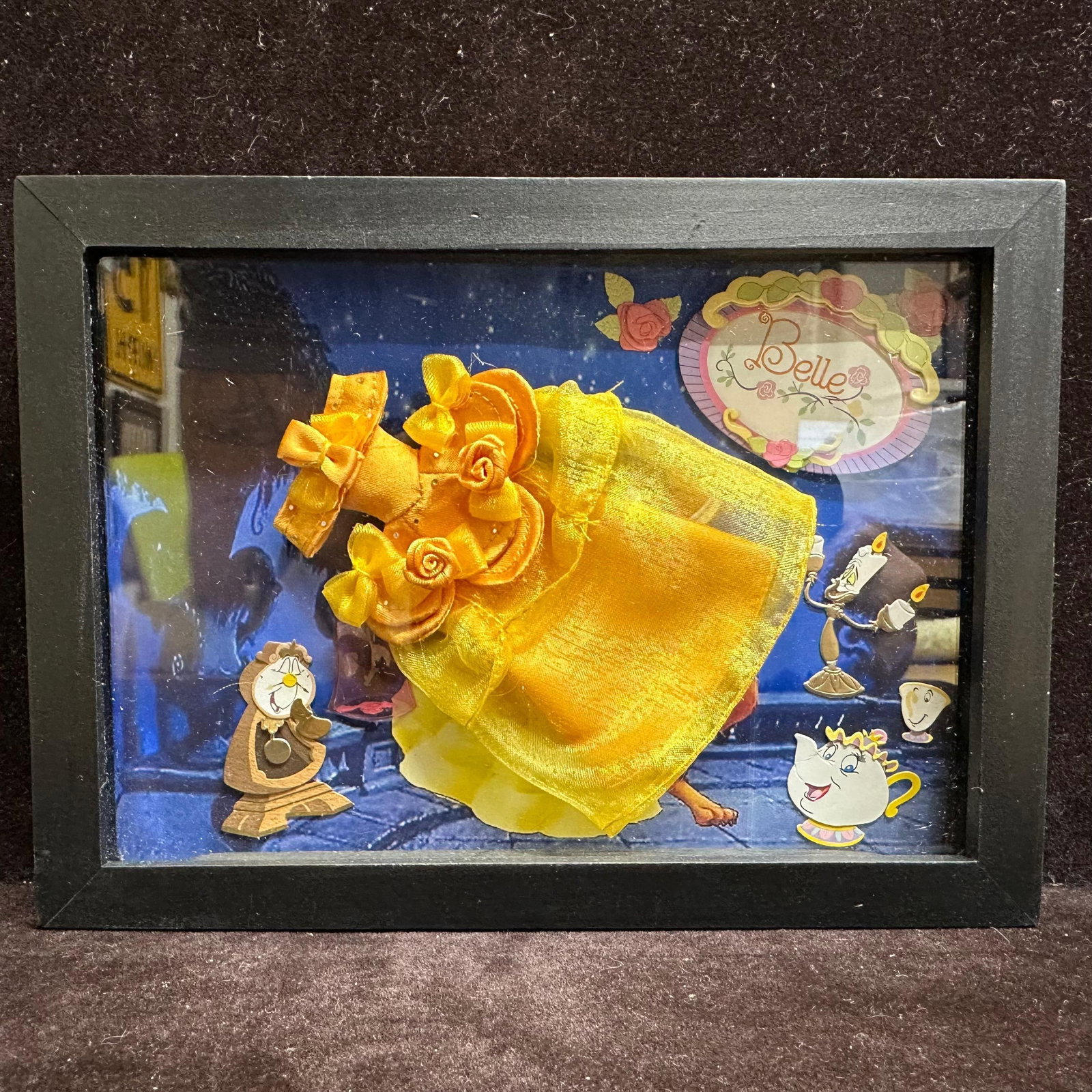 Beauty and the Beast Belle Yellow Doll Dress Shadowbox Display: Cute small black shadowbox featuring Belle’s iconic golden ballgown displayed in a three-dimensional presentation. The background features classic Beauty and the Beast characters, including Cogs