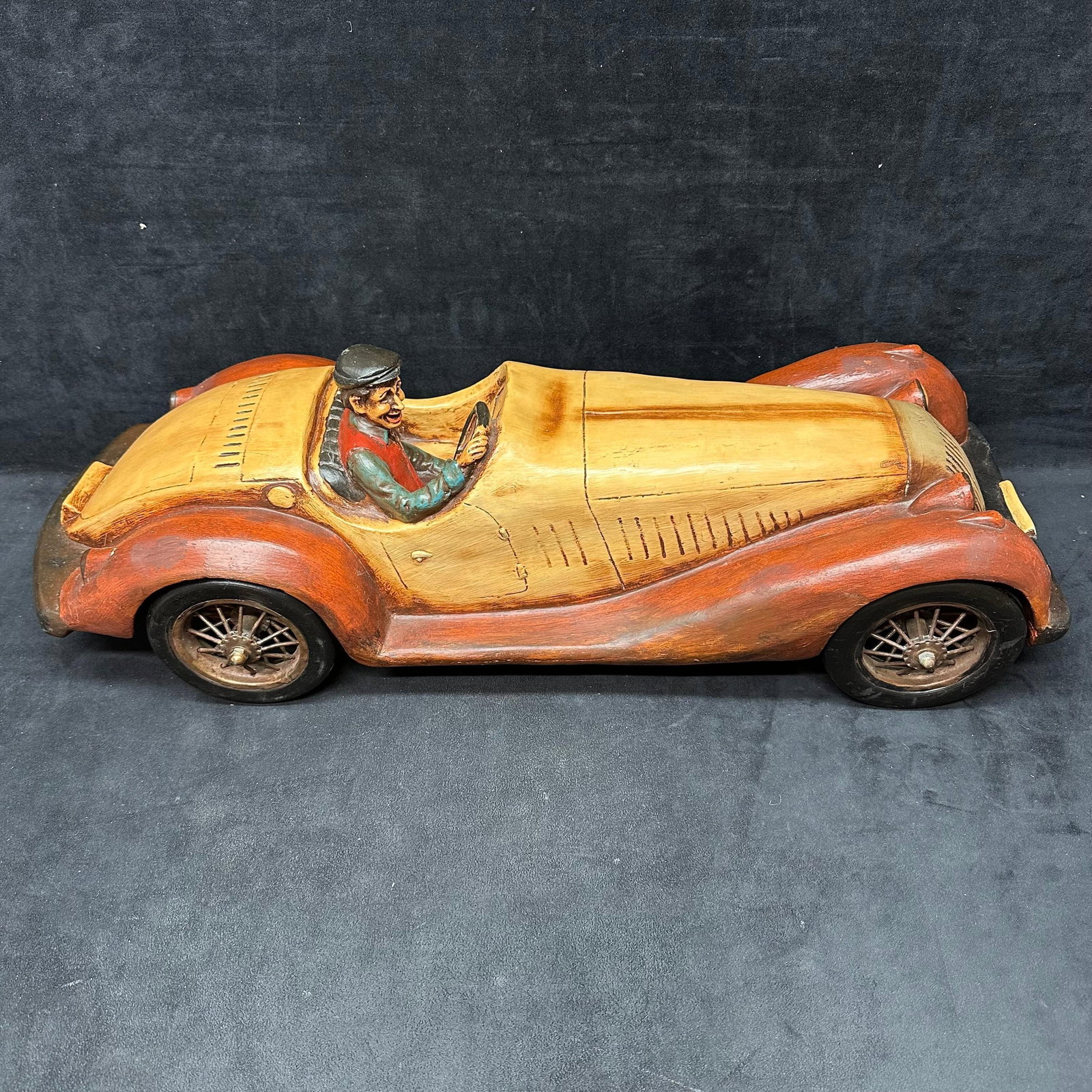 Resin Wooden 1934 Roadster Sculpture with Driver Figure Folk Art Style: Hand-carved wooden/resin sculpture depicting a classic open-top roadster with a seated driver figure. Carved and painted in warm natural wood tones with contrasting darker fenders, spoked wheels, and