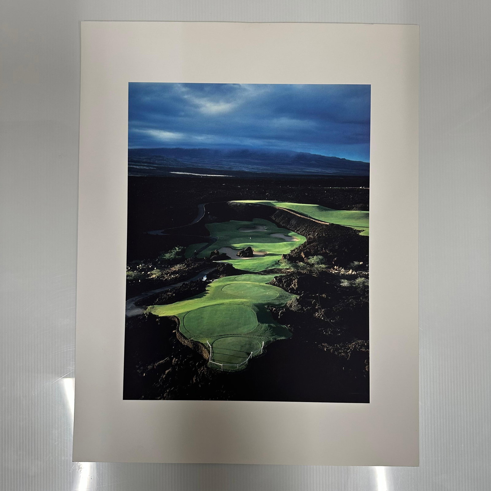 Aerial Golf Course Landscape Over Fairways Fine Art Color Photography Print: Vivid color photograph depicting an aerial view of a golf course set against dark rocky terrain and a dramatic twilight sky. The striking contrast of emerald greens and deep blues captures the serene 