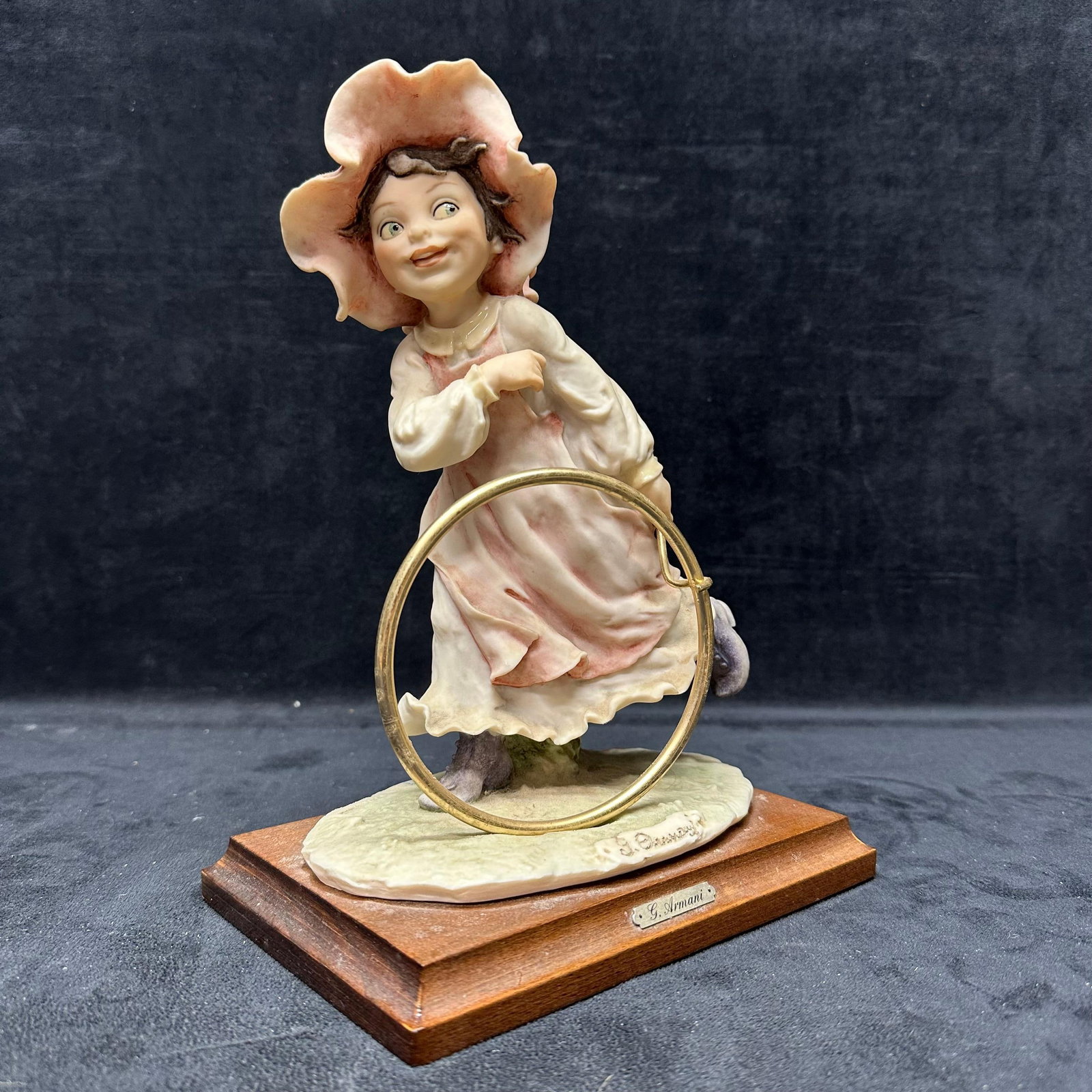 G. Armani Girl with Hoop Figural Sculpture Wooden Base: G. Armani sculpture depicting a young girl running with a brass hoop, wearing a flowing dress and a large bonnet. Hand-finished composition with soft pastel tones, mounted on a wooden display base.