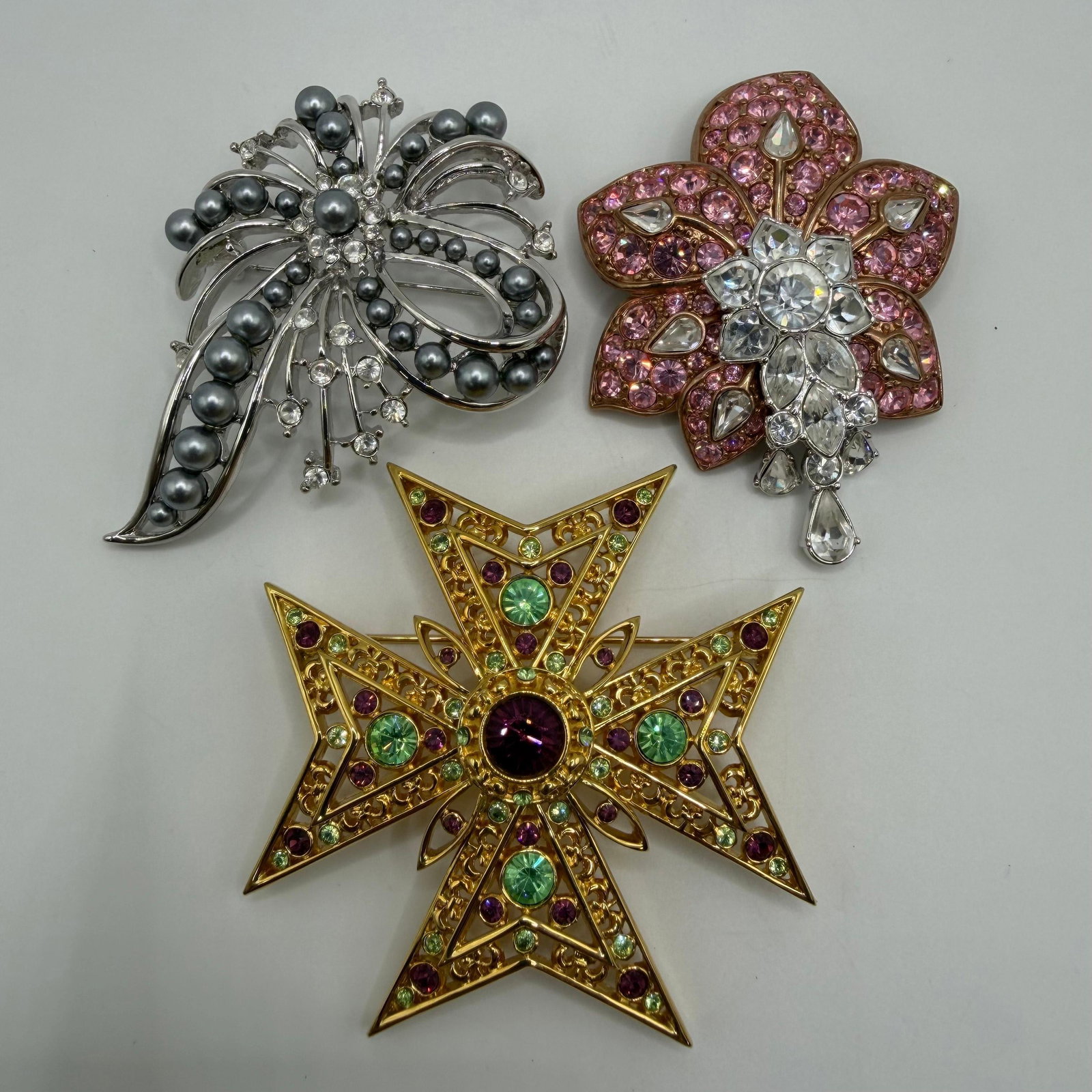 Three Designer Nolan Miller Rhinestone Statement Brooches AB5: Lot of three signed Nolan Miller designer brooches. Includes a silver-tone floral spray brooch with gray faux pearls and clear rhinestones, a rose-gold-tone pink rhinestone flower brooch with clear cr