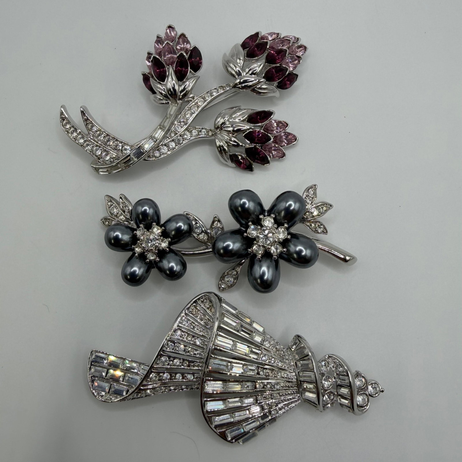 Designer Nolan Miller Crystal Rhinestone Silver Tone Brooches Floral and Ribbon Motif: Collection of three signed Nolan Miller silver-tone designer brooches. Includes a floral spray brooch accented with purple marquise-cut crystals and clear rhinestones, a double flower brooch featuring