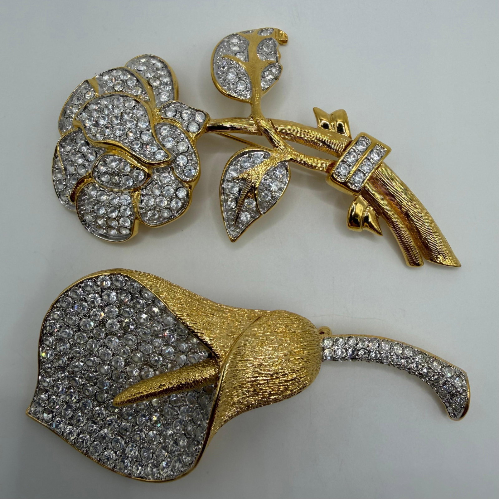 Nolan Miller Designer Crystal Floral Gold Tone Brooches Rose and Calla Lily Brooch Pins: Lot of two signed Nolan Miller designer brooches, including an elegant rose bouquet and a large calla lily. Both feature gold-tone settings with clear pave rhinestones throughout and secure locki